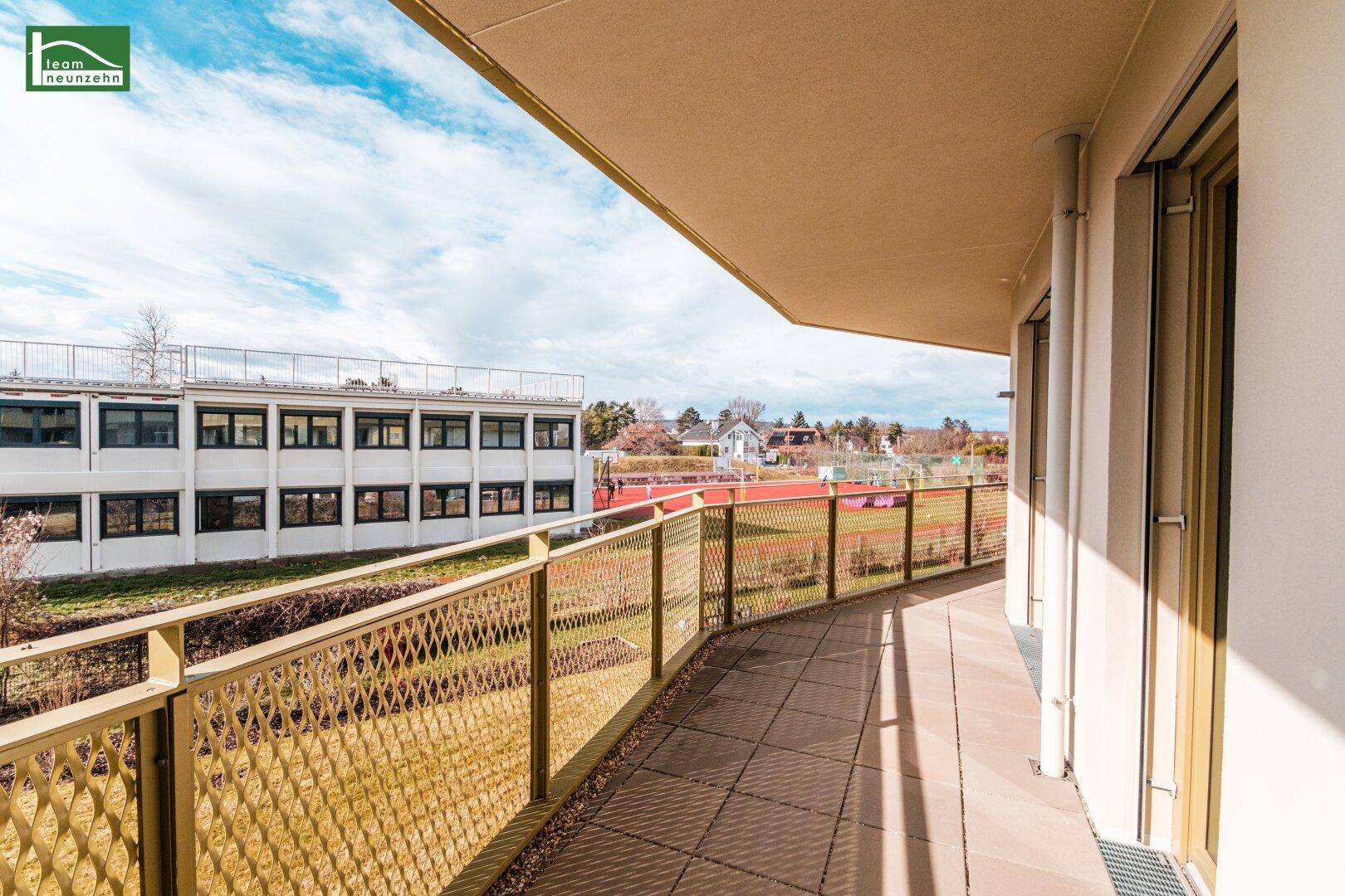 Langer Balkon mit Fliesenboden und Blick auf eine Grünfläche und Sportanlagen.