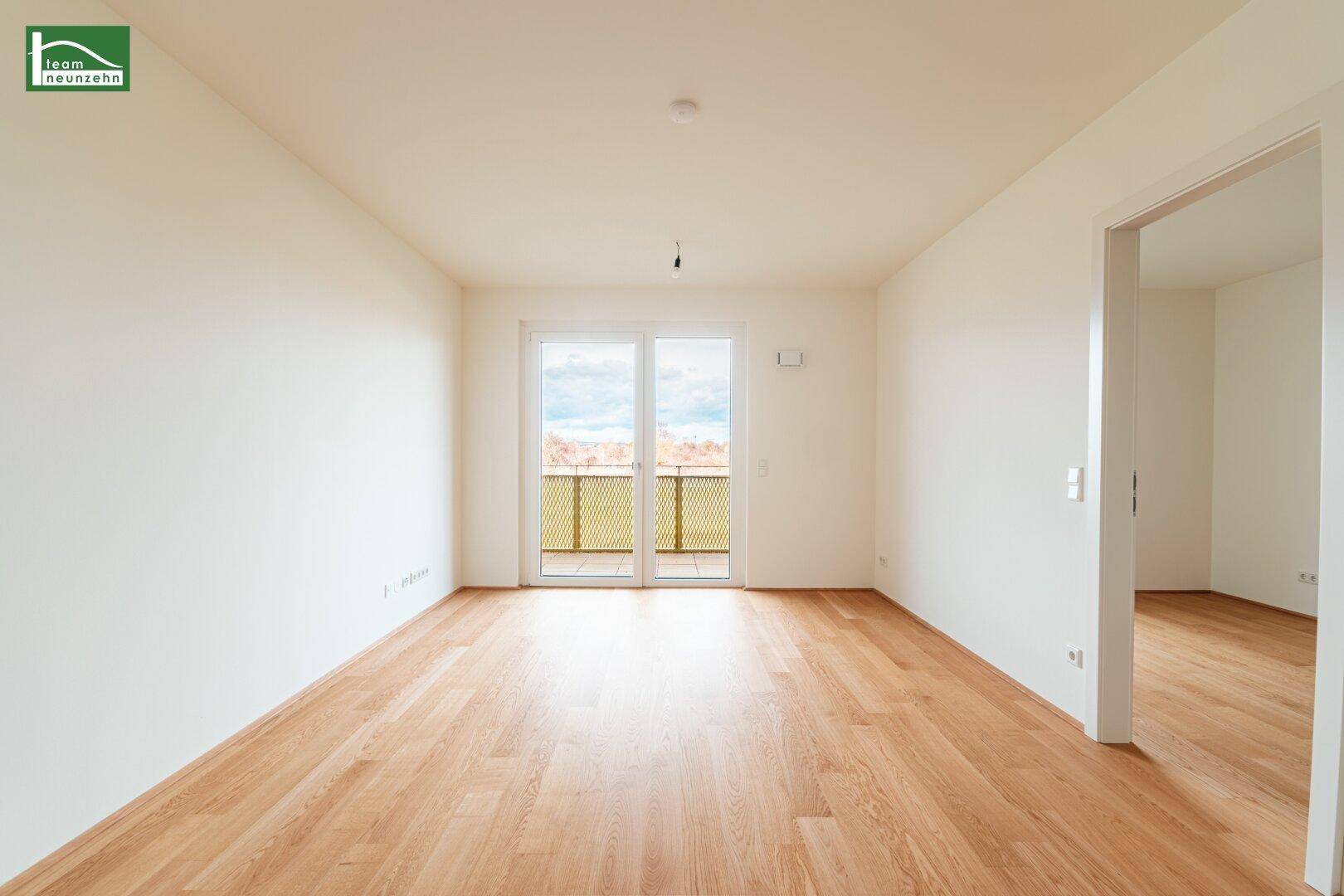 Helles, leeres Zimmer mit hellem Holzfußboden und Zugang zu einem Balkon.