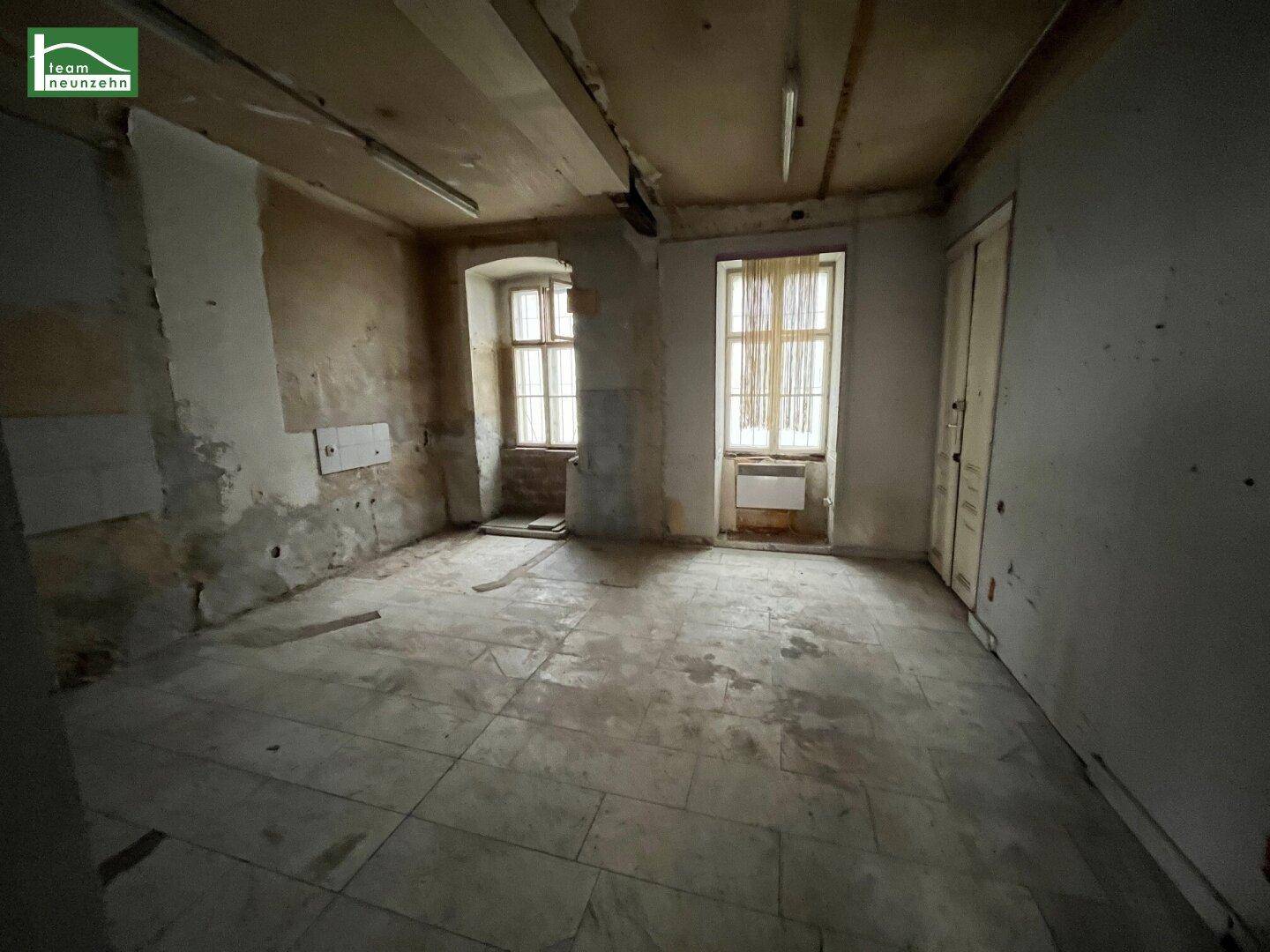 Leerer Raum mit zwei Fenstern, Fliesenboden und sichtbaren Renovierungsarbeiten an den Wänden.