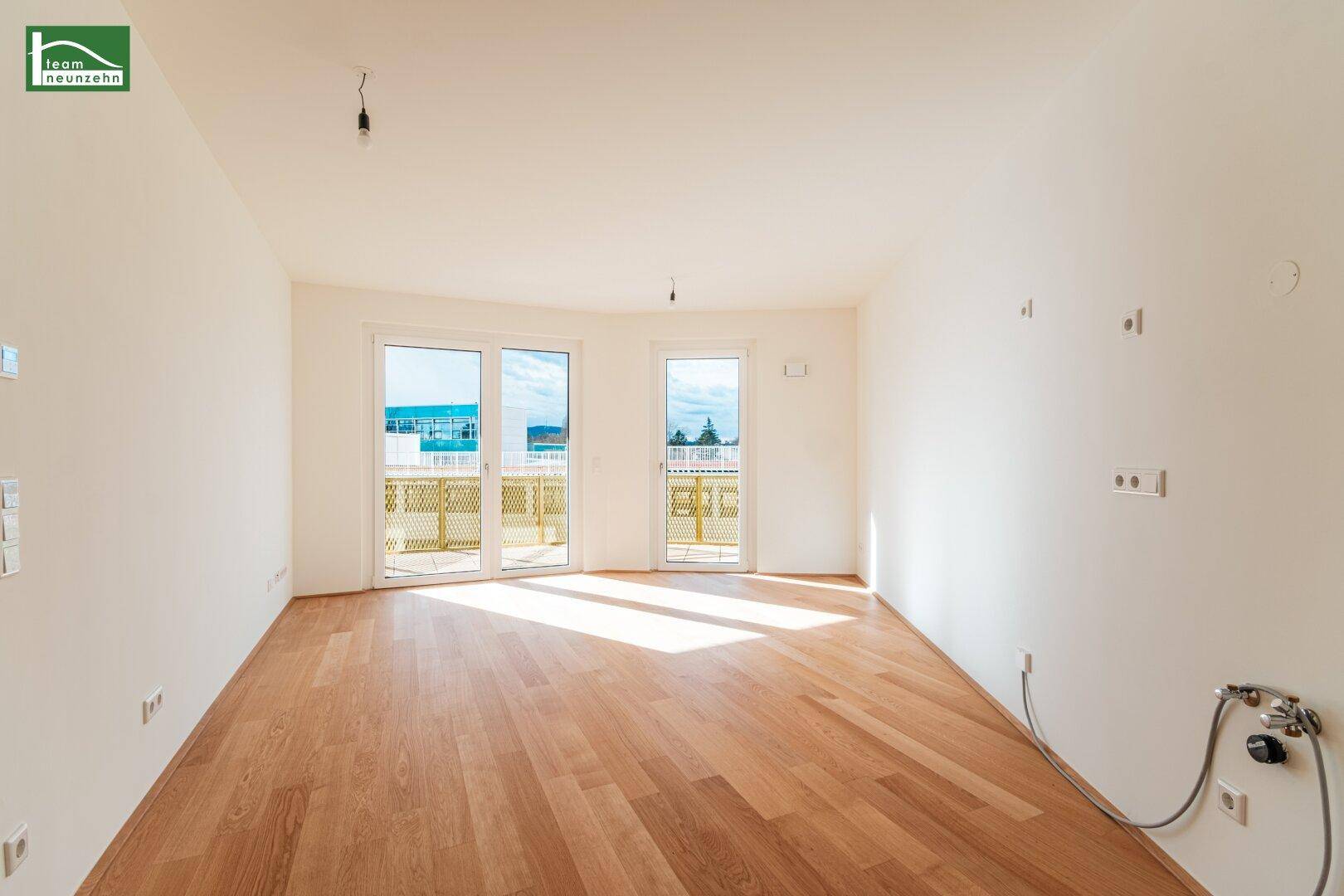 Heller, leerer Wohnraum mit Holzboden und Zugang zu einem sonnigen Balkon.