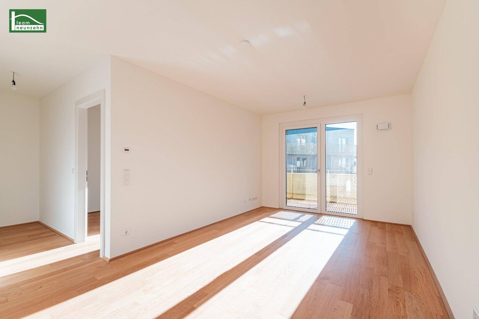 Helles, leeres Zimmer mit Holzboden und Zugang zu einem Balkon, durchflutet von Sonnenlicht.