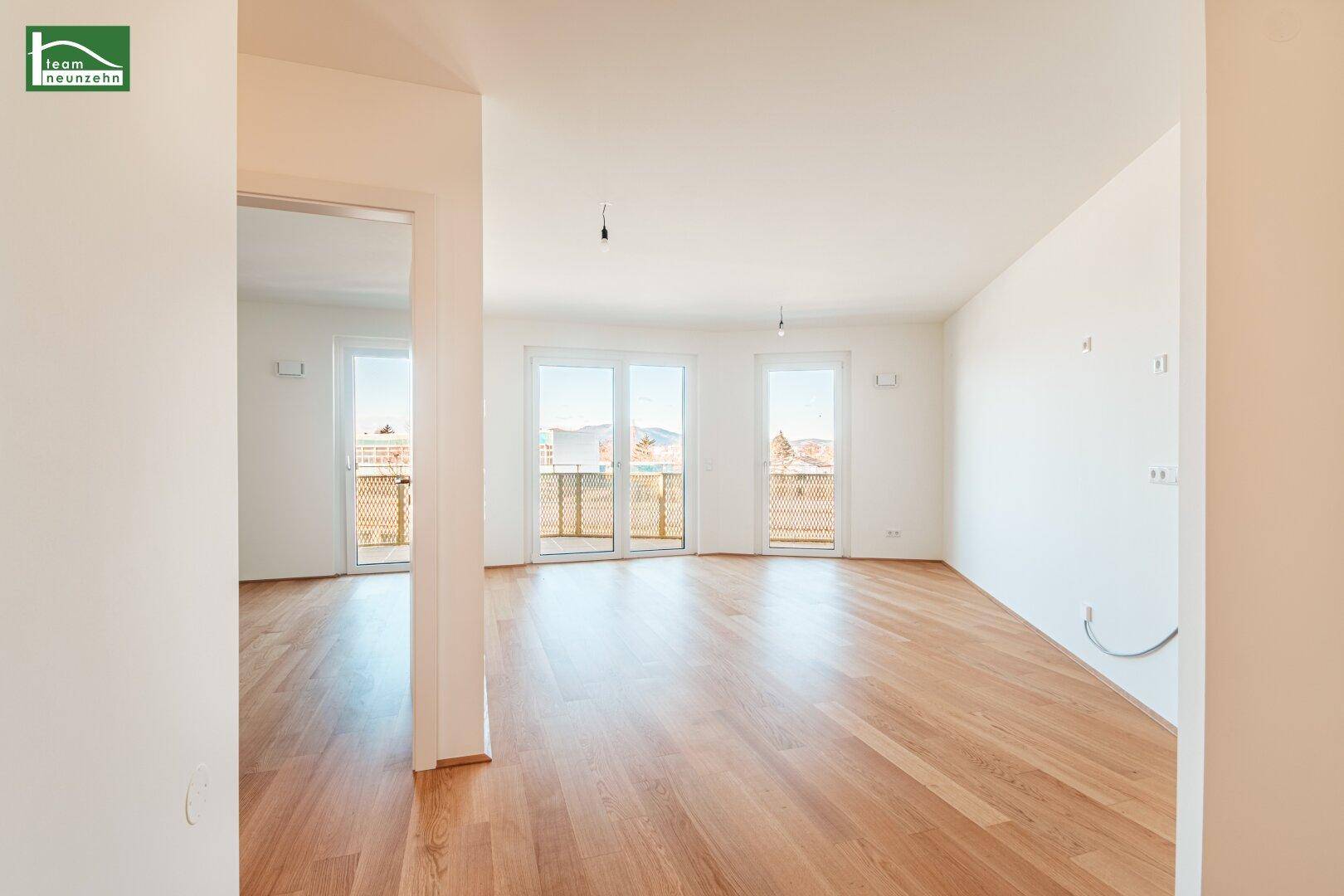 Heller, leerer Wohnraum mit Holzboden und großen Fenstern, die Zugang zu einem Balkon bieten.
