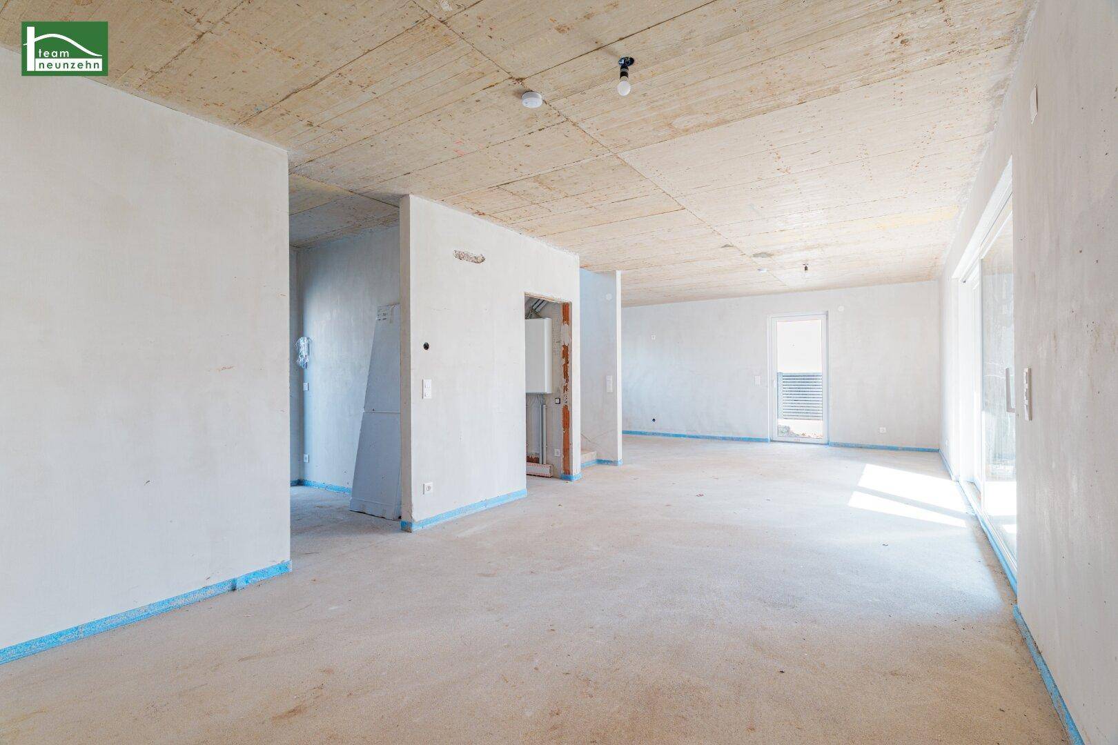 Großer, offener Innenraum im Rohbau mit Betonboden und viel Tageslicht durch Fenster.