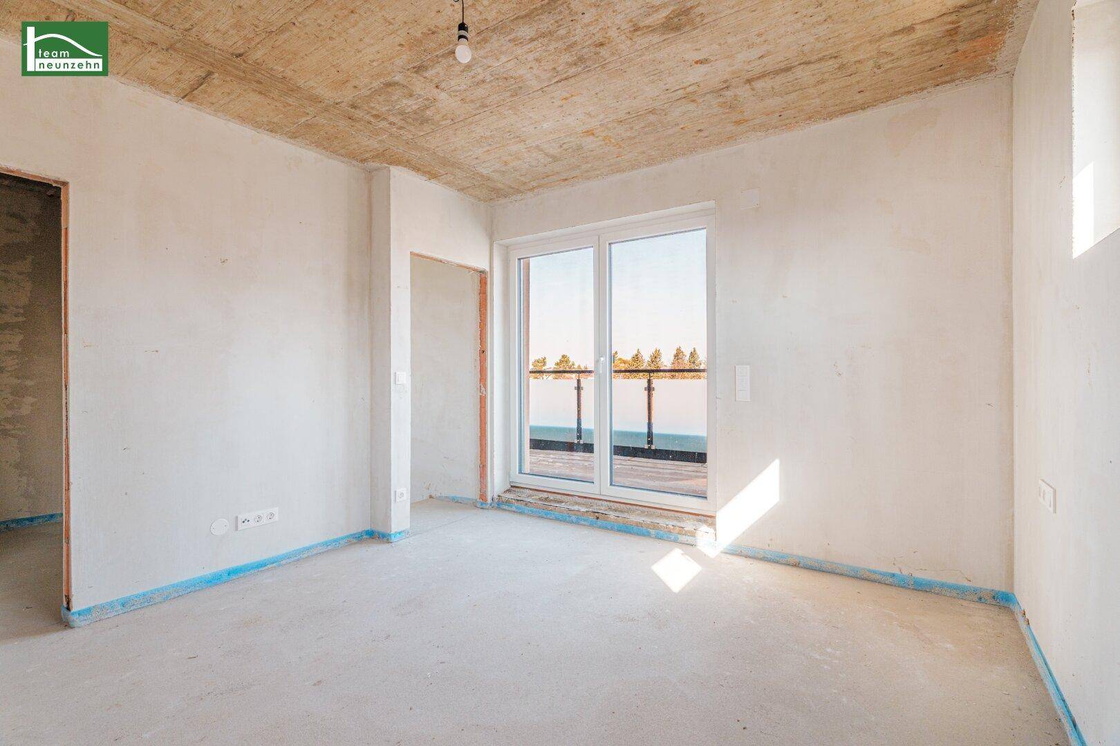 Heller Rohbau-Raum mit großem Fenster und Zugang zu einem Balkon, viel Tageslicht.