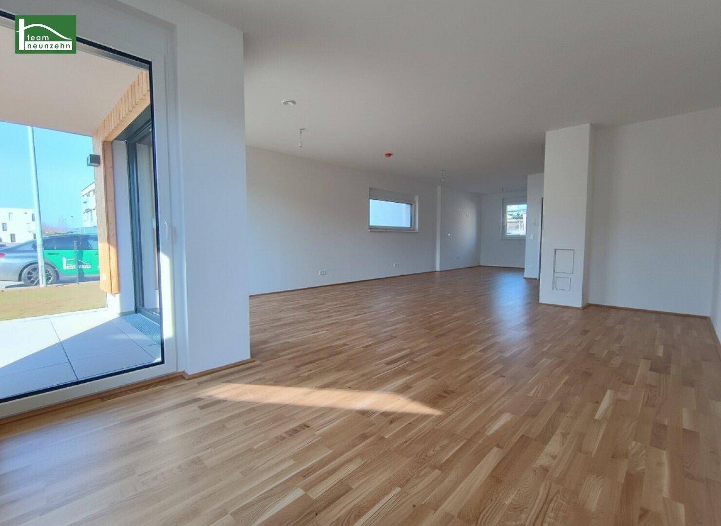Geräumiger, leerer Wohnbereich mit hellem Parkettboden und großem Fenster mit Zugang nach draußen.
