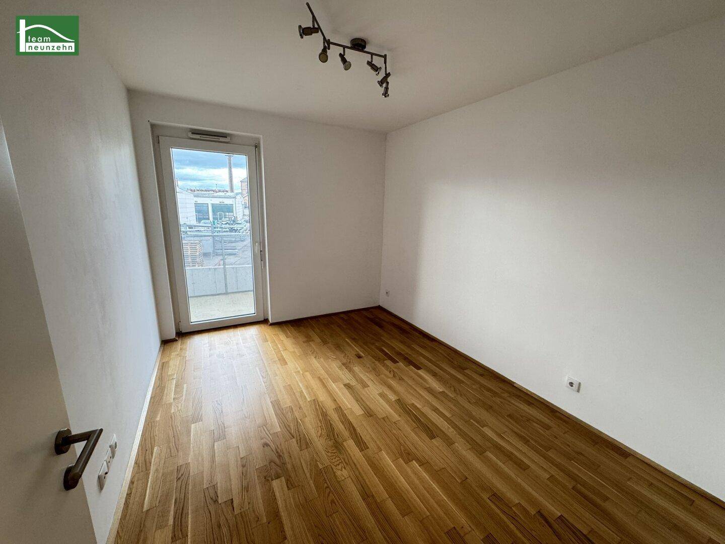 Helles, leeres Zimmer mit Parkettboden und Zugang zu einem Balkon.