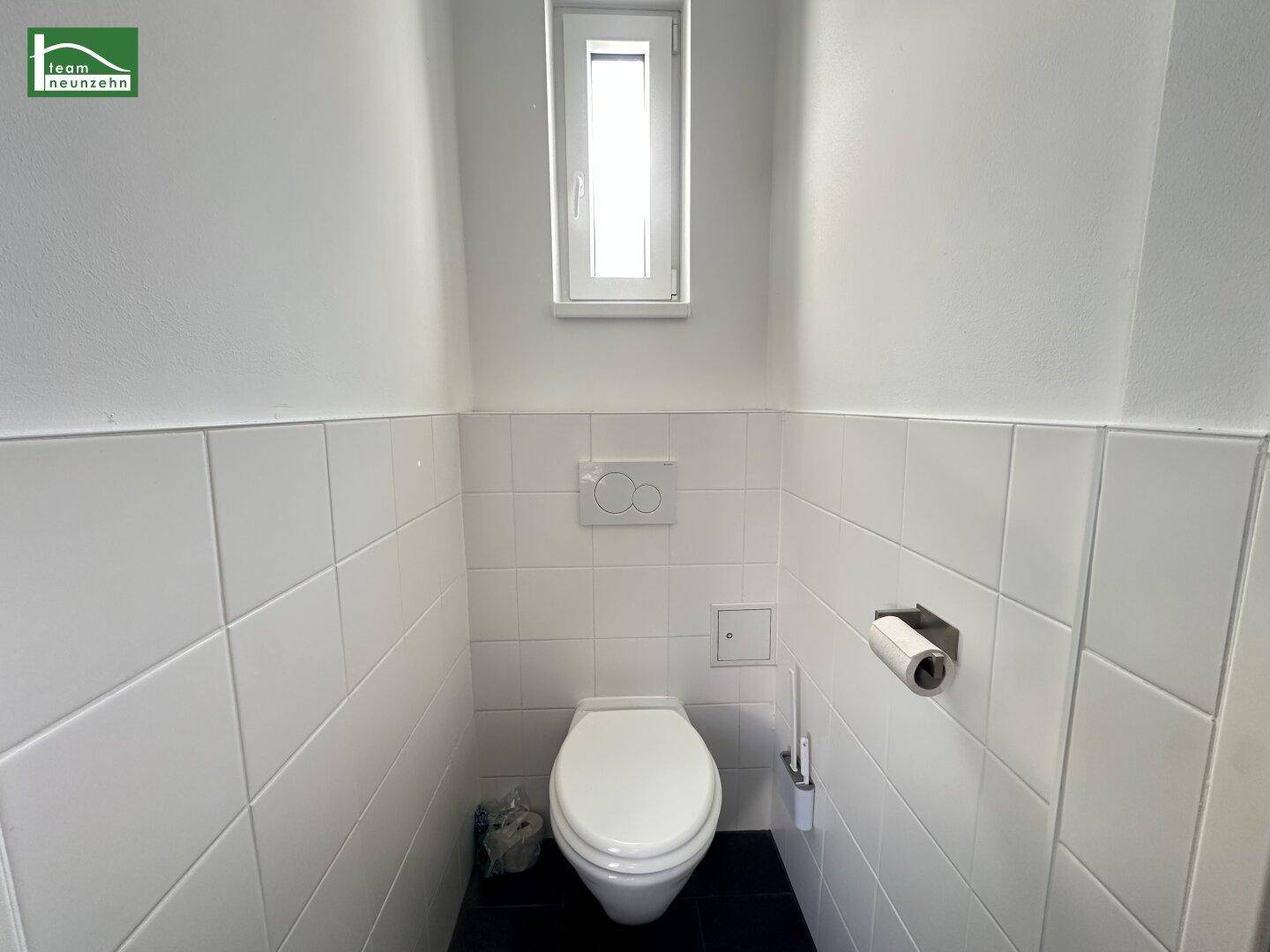 Separates WC mit wandmontierter Toilette, kleinem Fenster und weißen Wandfliesen.