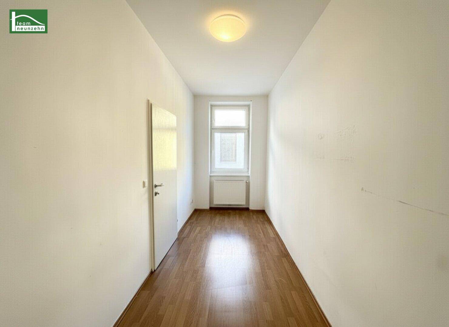 Schmaler Flur mit Laminatboden und einem Fenster am Ende, das Tageslicht hereinlässt.