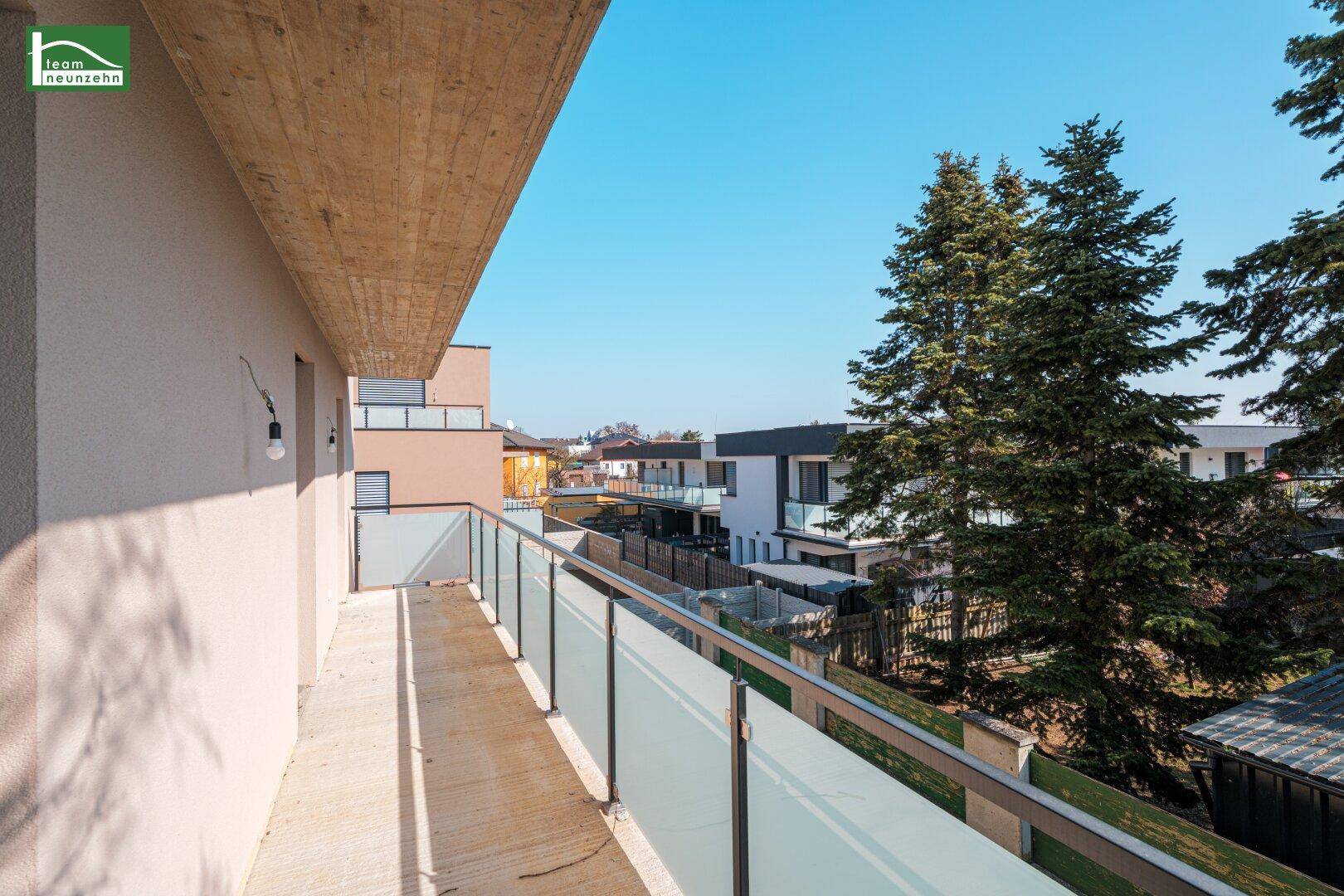 Langer Balkon mit Glasgeländer und Blick auf Bäume und die angrenzenden Häuser.