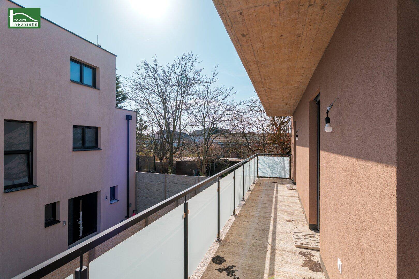 Geräumiger Balkon mit Glasgeländer und Blick auf Bäume und Nachbargebäude unter blauem Himmel.