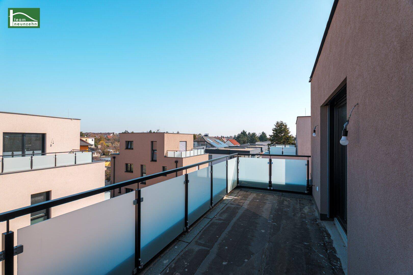 Breiter Balkon mit Glasgeländer und Blick auf die umliegenden Neubauten und Dächer.