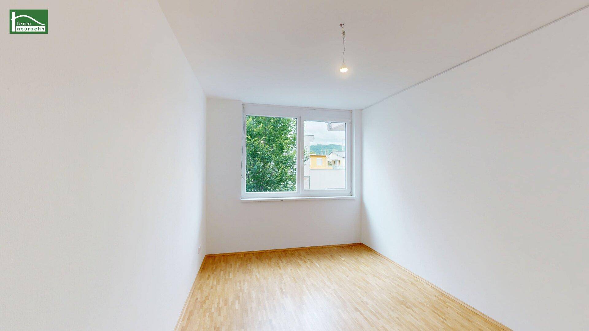Helles Schlafzimmer mit Parkettboden und Fenster, das einen Blick ins Grüne ermöglicht.