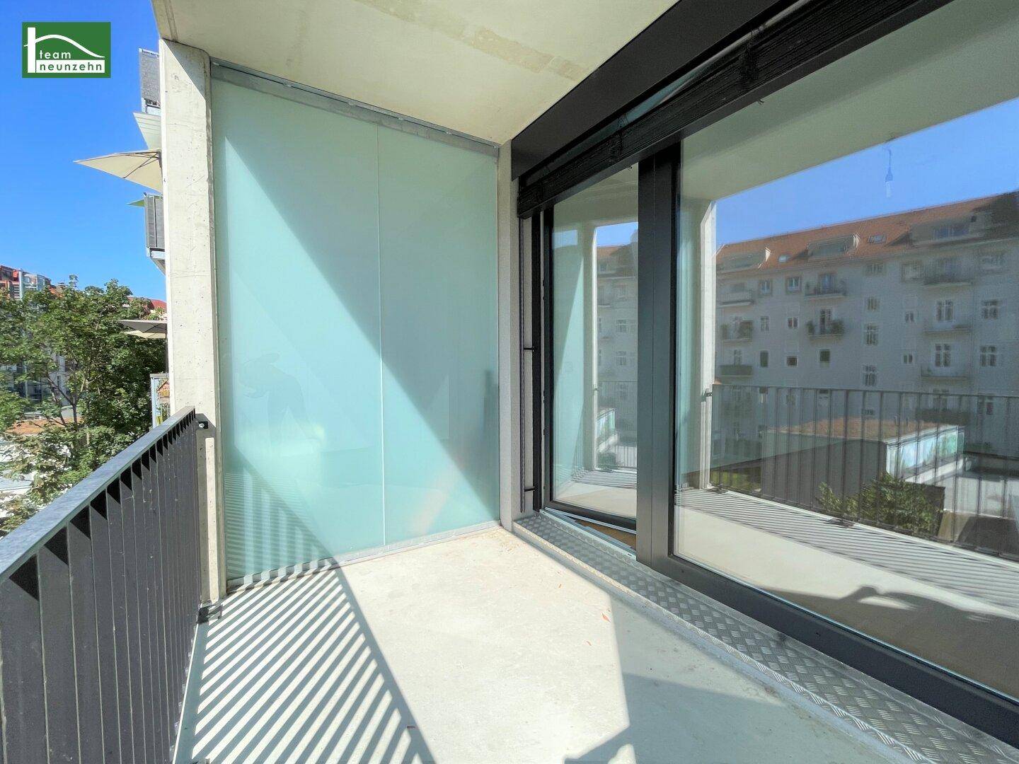 Moderner Balkon mit Betonboden und Glasbrüstung, bietet Blick auf die umliegenden Gebäude.