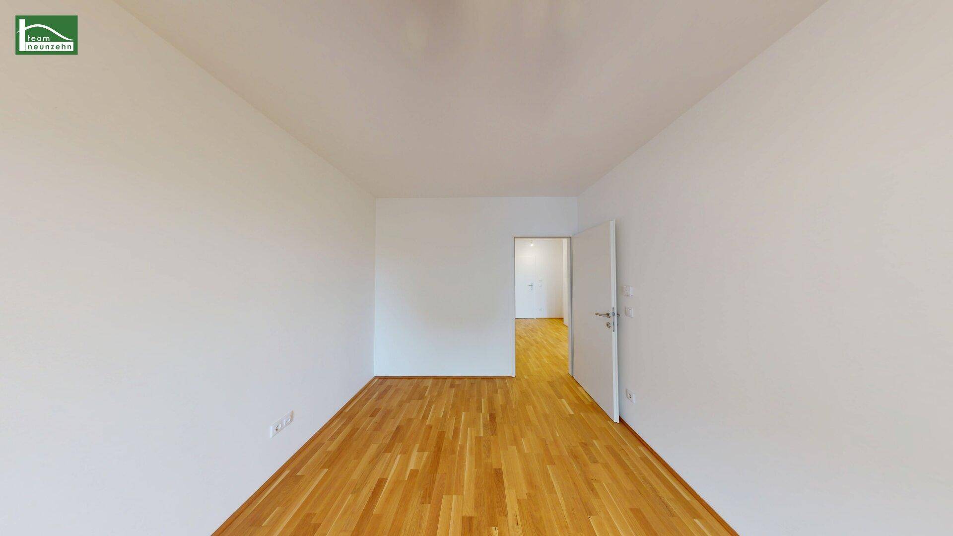 Heller, leerer Raum mit Holzboden und offener Tür zu einem angrenzenden Bereich.