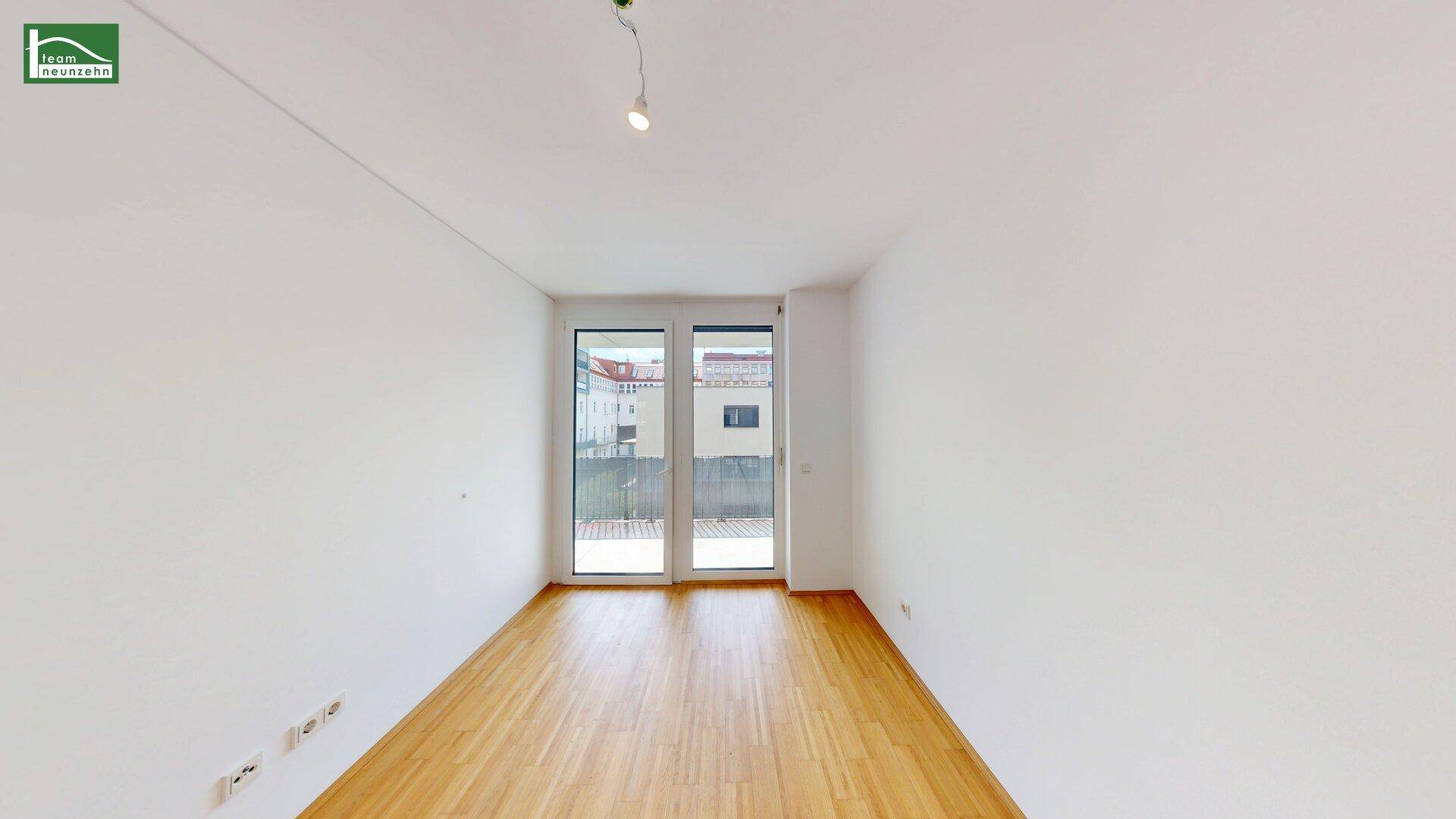 Heller, leerer Raum mit Parkettboden und großen Fenstertüren zum Balkon.