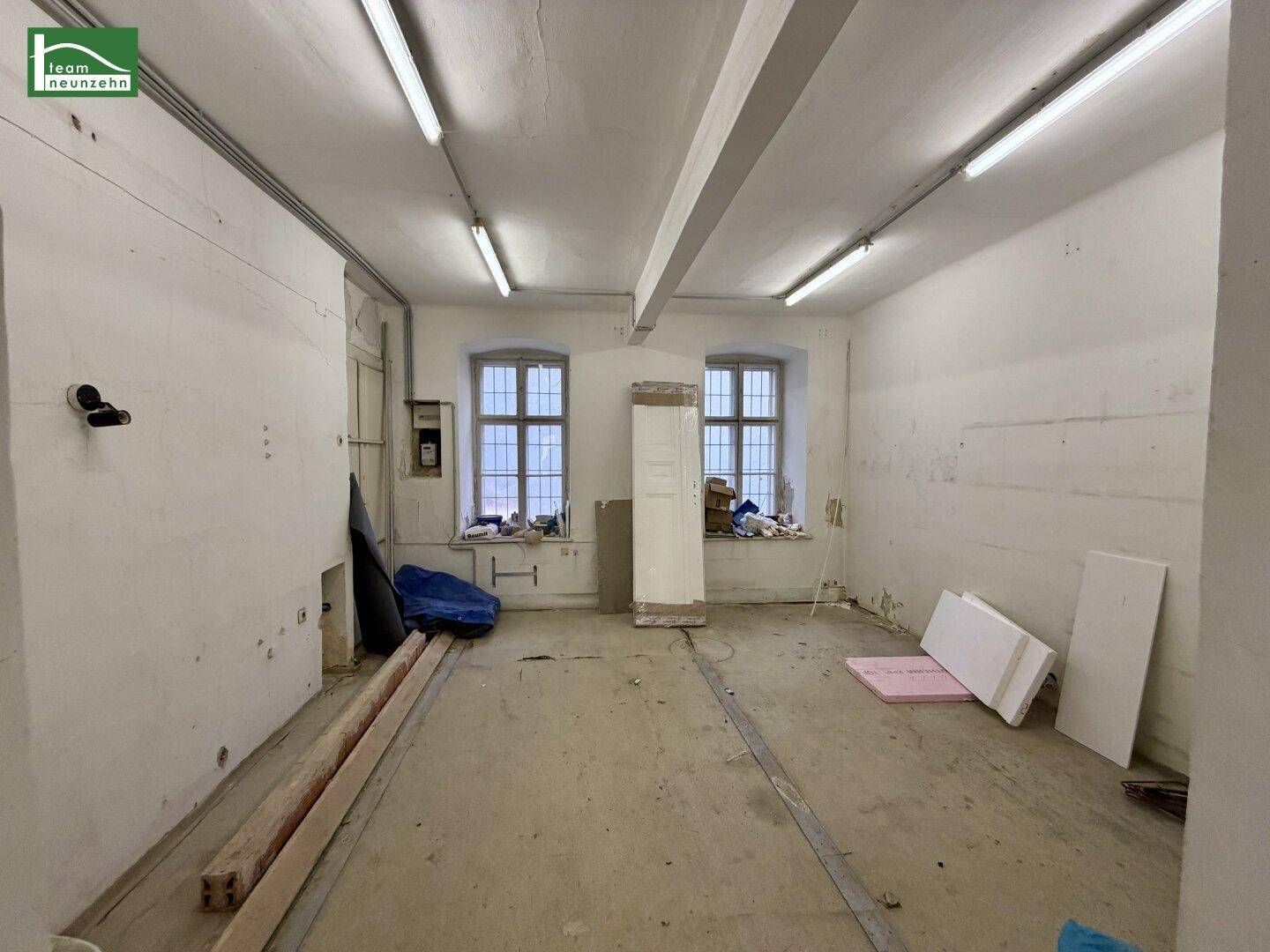 Großer, leerer Raum mit zwei Fenstern und einfacher Beleuchtung, bereit für Renovierung.