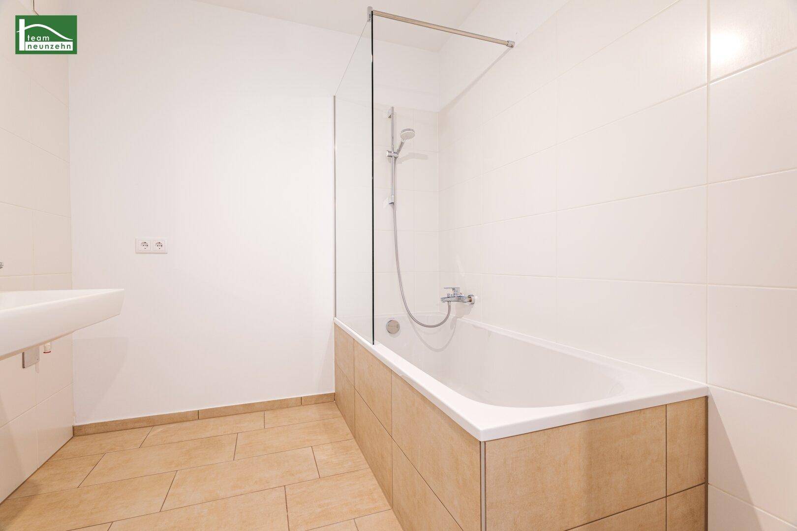 Helles Badezimmer mit Badewanne, Duschvorrichtung und beigefarbenen Bodenfliesen.