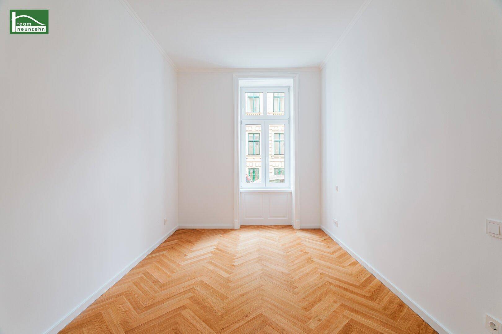 Helles Zimmer mit Fischgrätparkett und einem hohen Fenster mit Blick nach draußen.