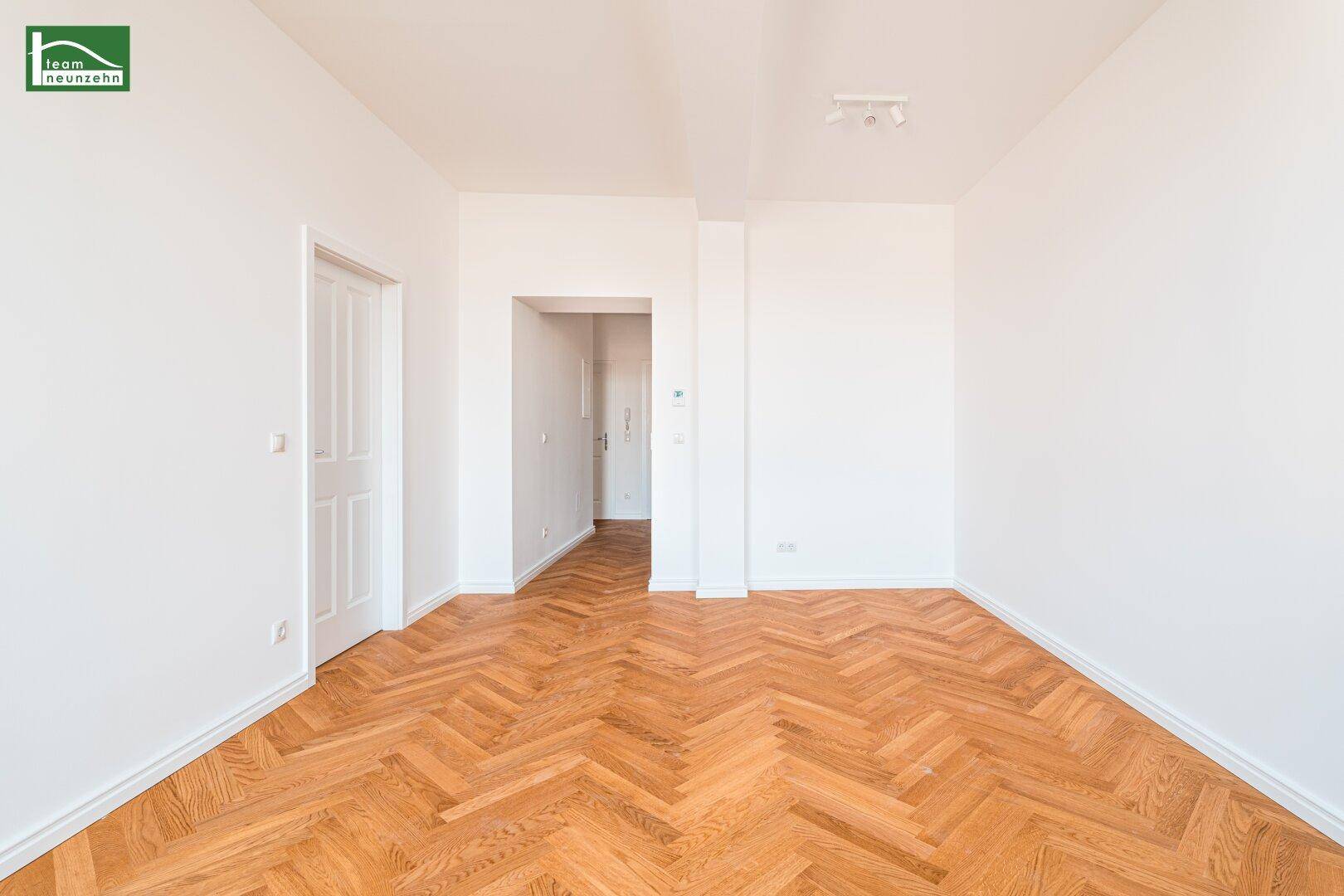 Heller, leerer Raum mit Fischgrätparkettboden und weißen Wänden, Blick in den Flur.