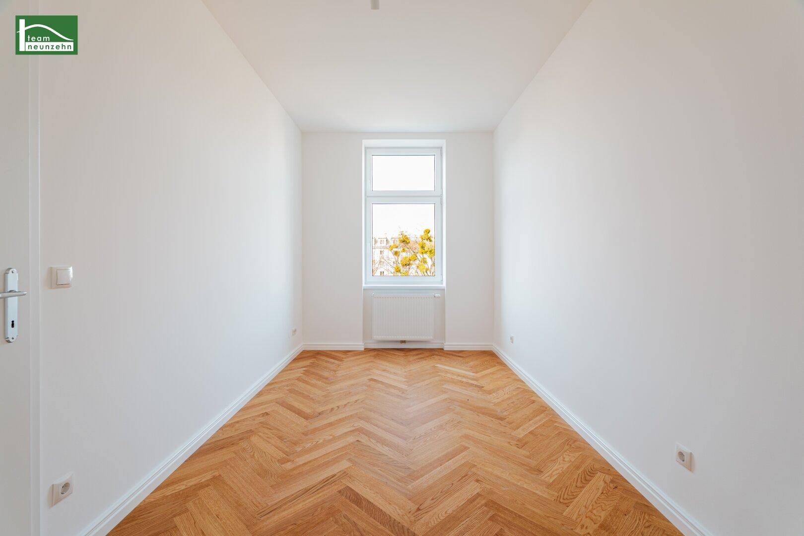 Schmaler, heller Raum mit einem Fenster, Fischgrätparkett und weißen Wänden.