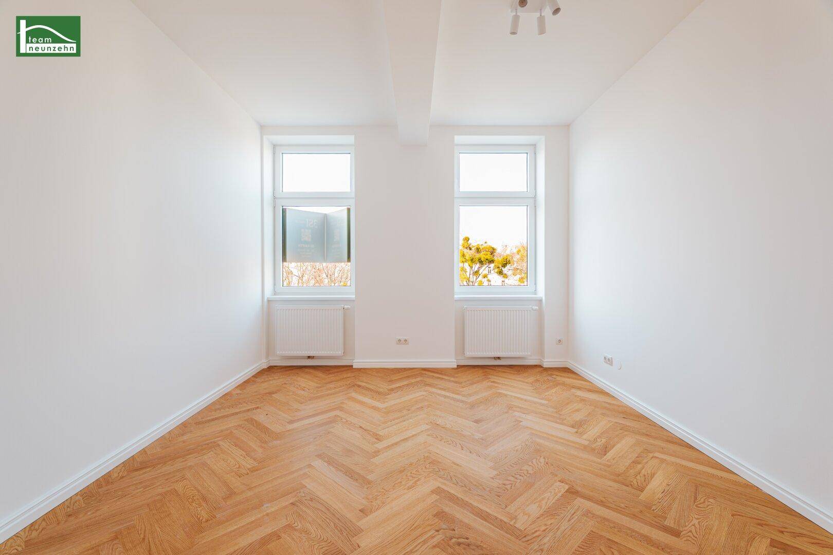 Lichtdurchfluteter, leerer Raum mit zwei Fenstern, Fischgrätparkett und weißen Wänden.