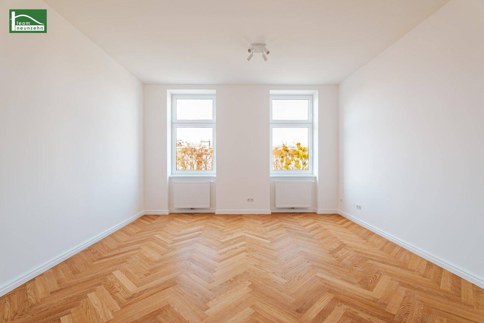 Lichtdurchflutetes Zimmer mit zwei Fenstern, Heizkörpern und klassischem Fischgrätparkett.