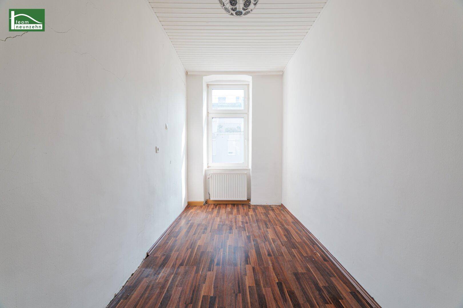 Schmaler Flur mit dunklem Laminatboden und einem Fenster am Ende des Raumes.