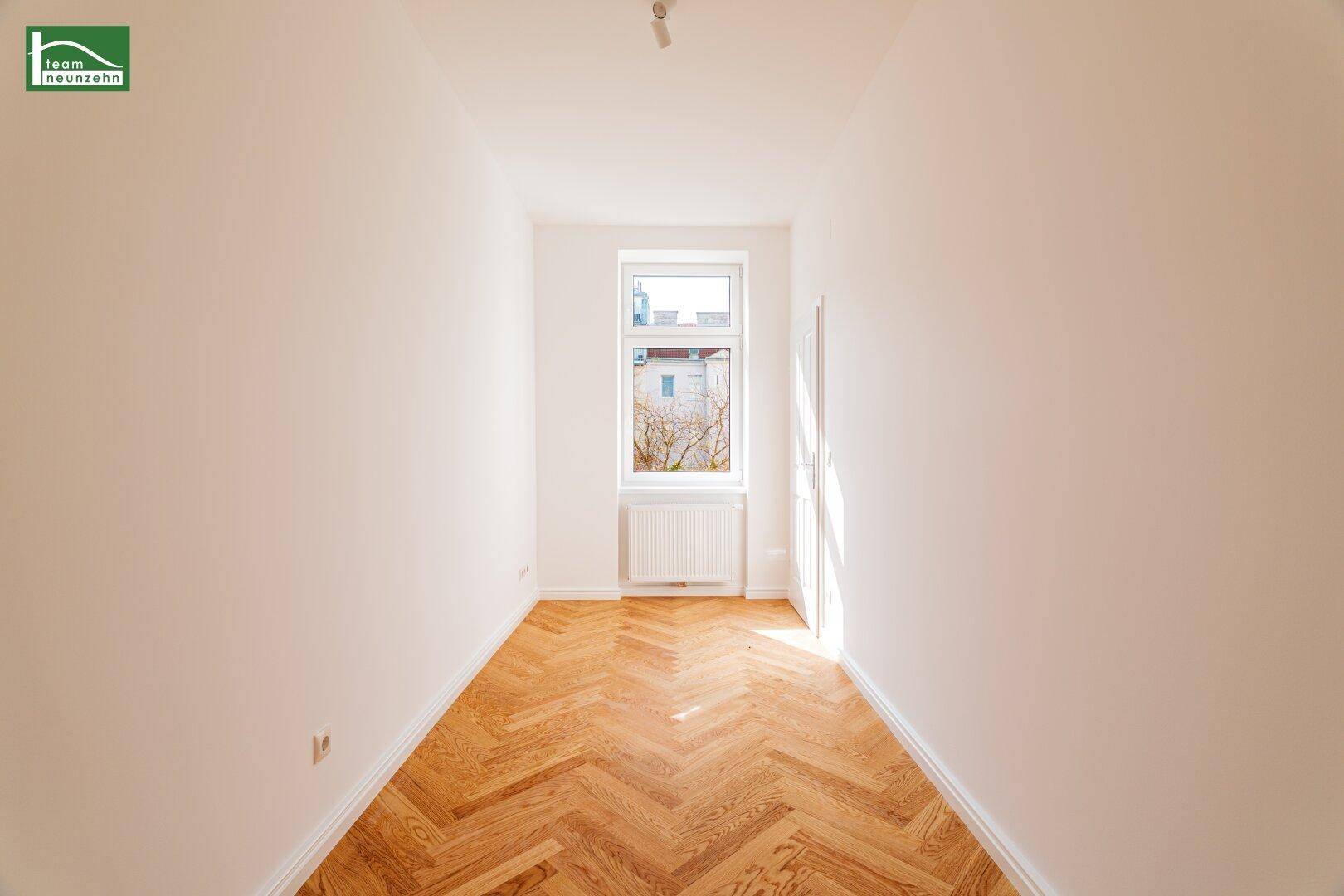 Schmaler, heller Raum mit Fischgrätparkett und einem Fenster, das Tageslicht spendet.