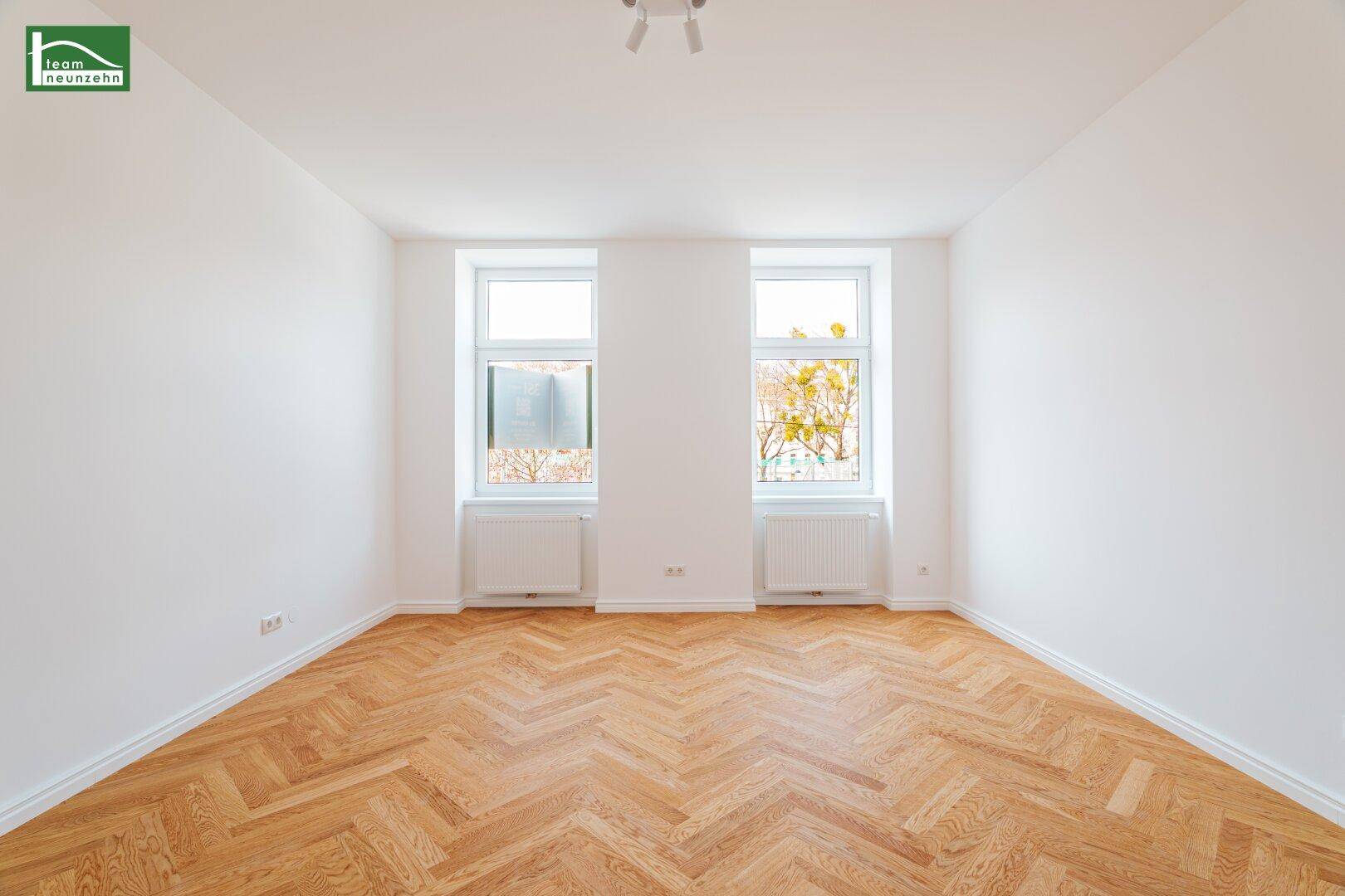 Lichtdurchflutetes Zimmer mit zwei großen Fenstern, Fischgrätparkett und weißen Heizkörpern.