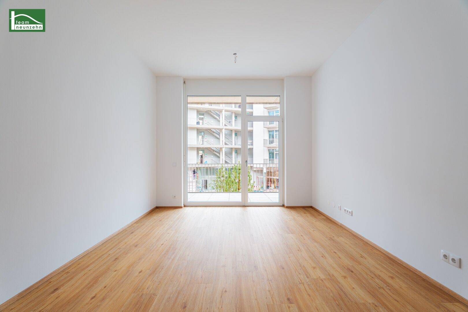 Heller Raum mit Holzboden und großem Fenster, das Zugang zu einem Balkon bietet.