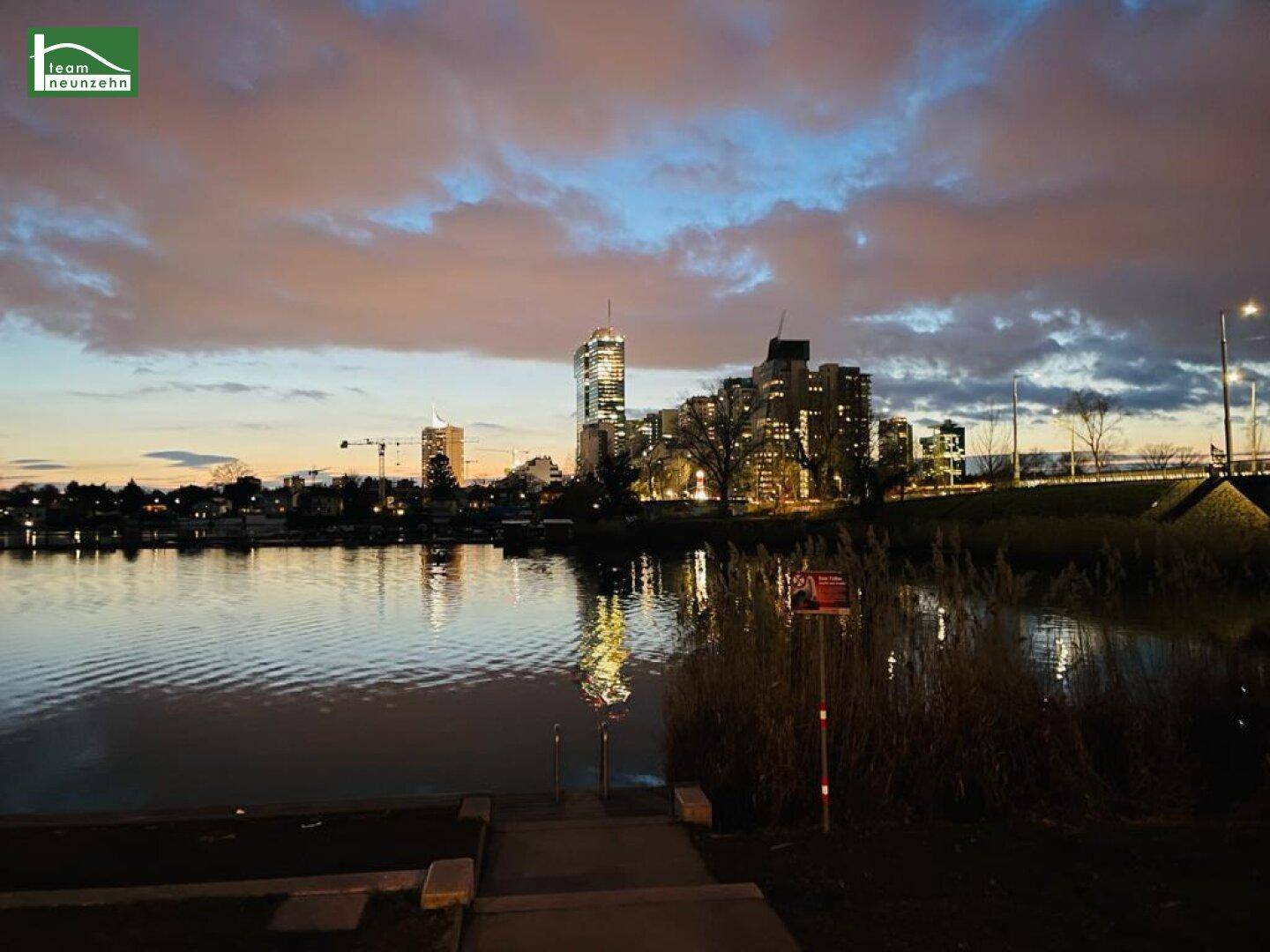 Abendlicher Blick auf die Stadt mit beleuchteten Gebäuden und einem Fluss im Vordergrund unter einem farbenfrohen Himmel.