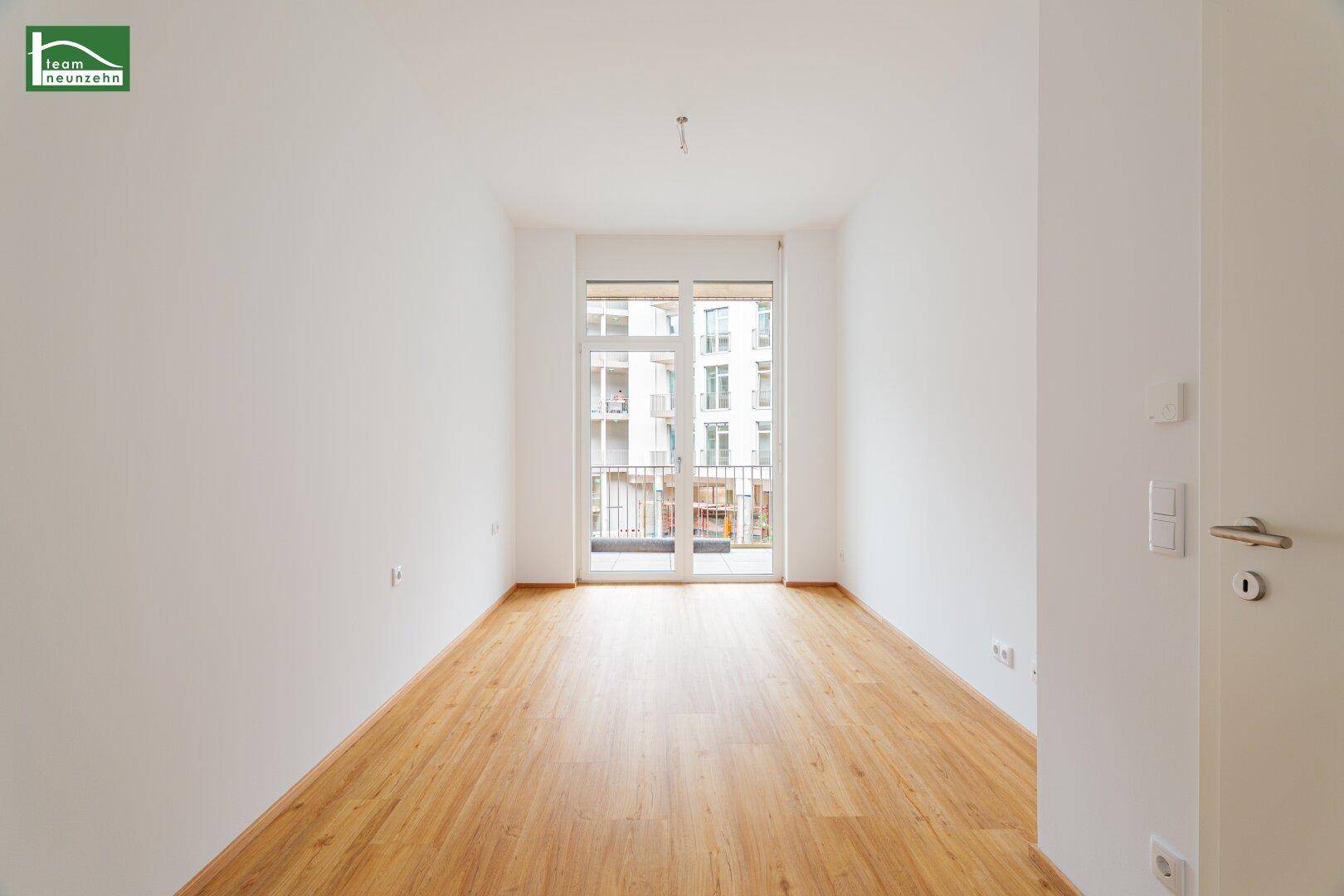 Leeres Zimmer mit hellem Holzfußboden und großen Fenstern, die zu einem französischen Balkon führen.
