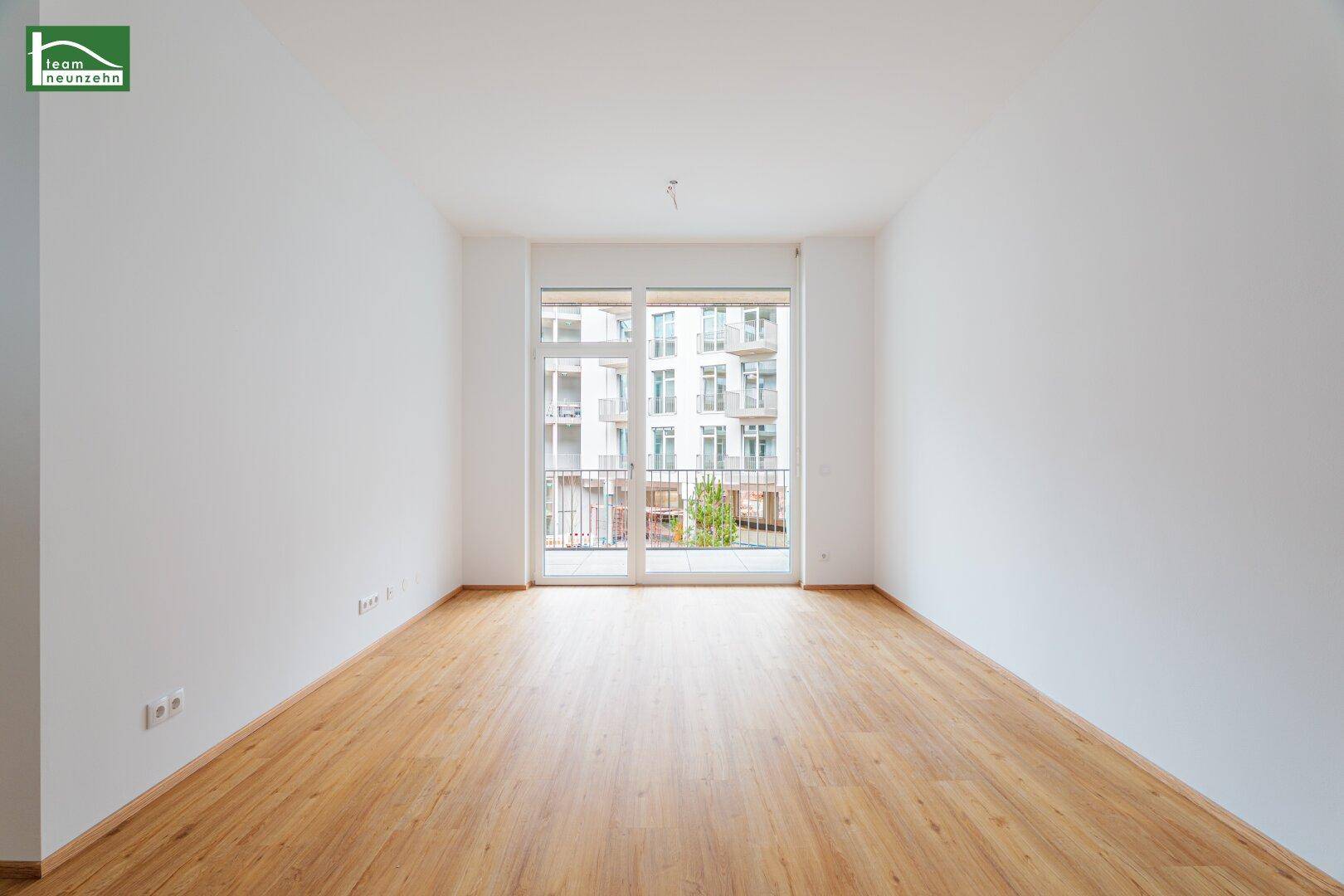 Leerer Wohnraum mit hellem Holzfußboden und Zugang zu einem französischen Balkon mit Blick auf Gebäude.