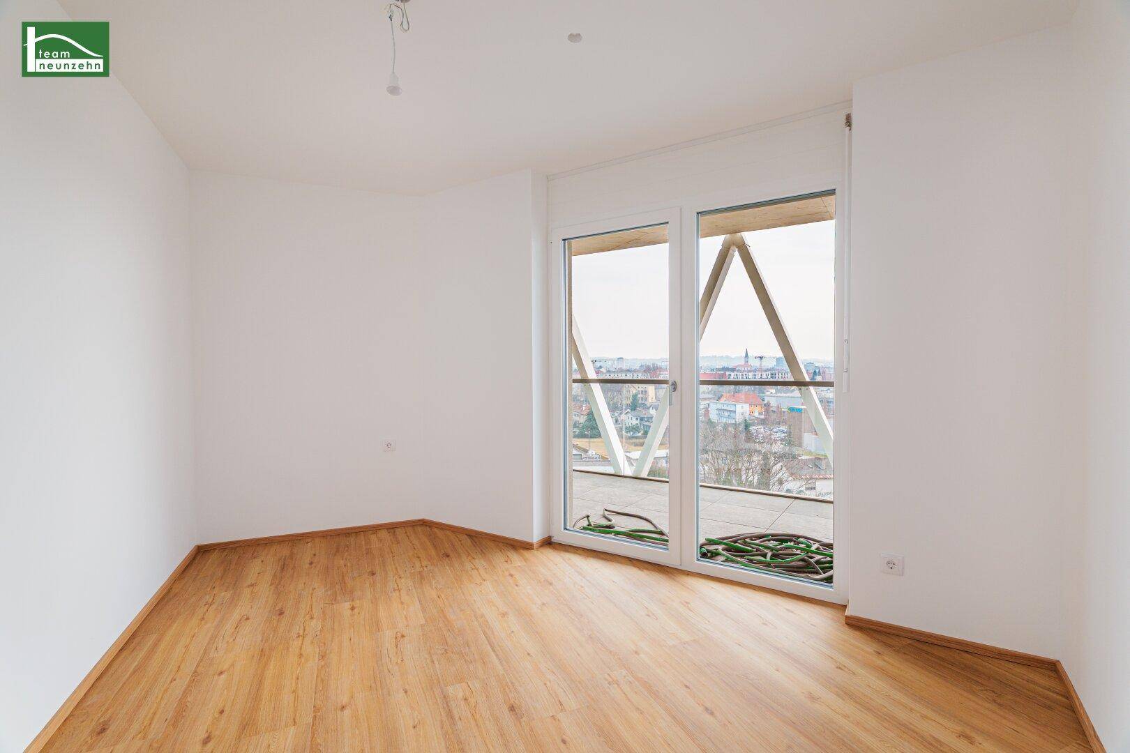 Leerer Raum mit Holzboden und großen Fenstern, die viel Tageslicht hereinlassen.