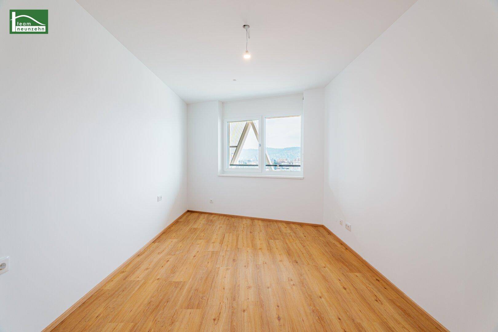 Leeres Zimmer mit Laminatboden, weißen Wänden und Fenster mit Blick auf die Umgebung.