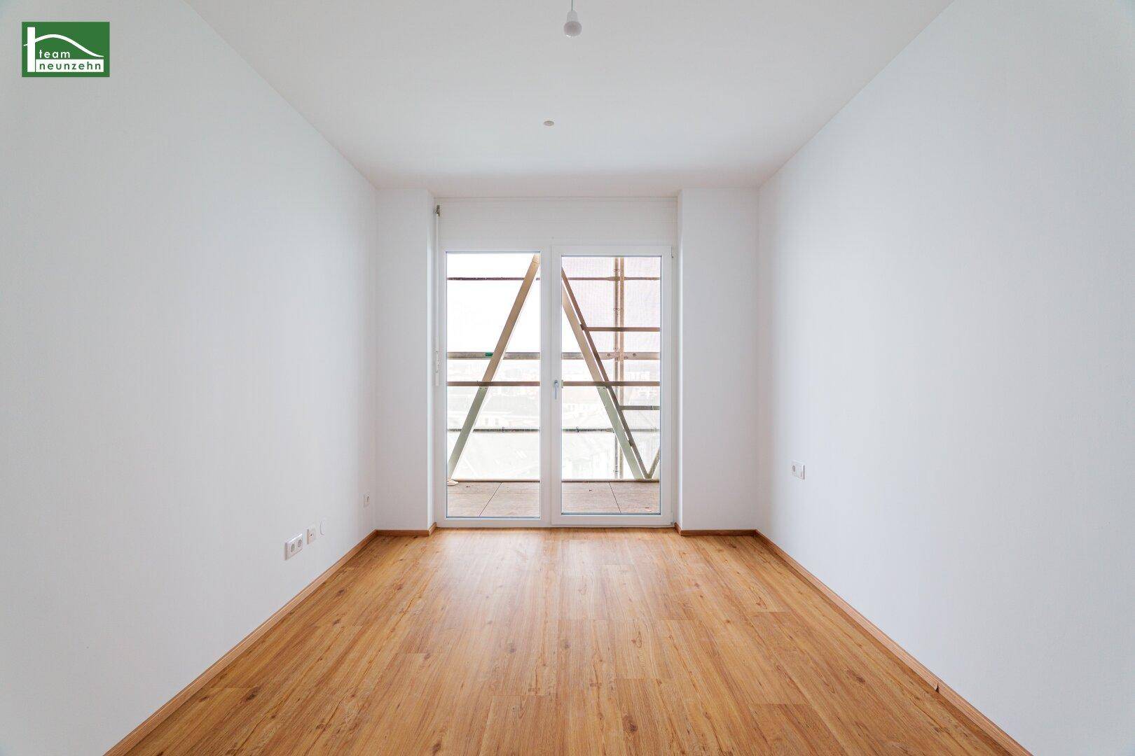 Geräumiges, unmöbliertes Zimmer mit Holzboden und großen Fenstern zum Balkon.