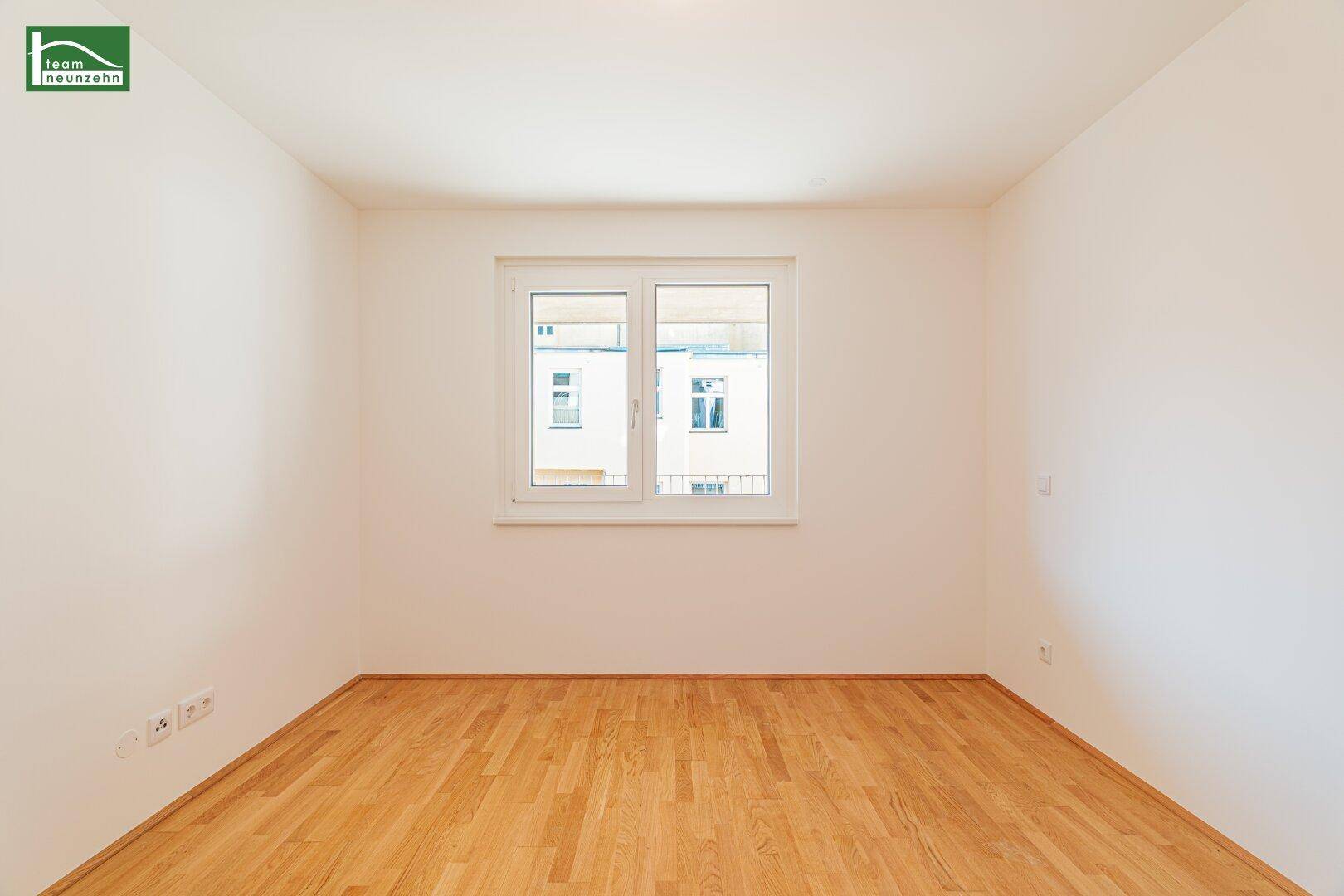 Leeres Schlafzimmer mit hellem Parkettboden und einem großen Fenster, das viel Tageslicht hereinlässt.