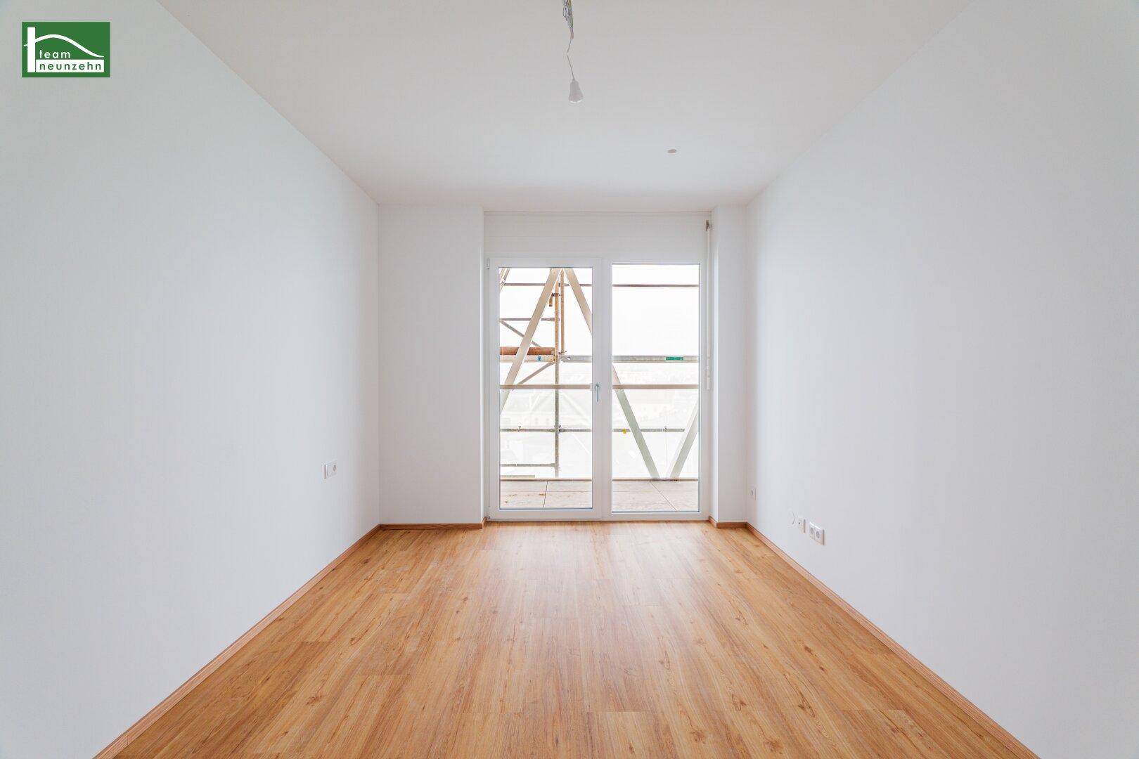 Leeres Zimmer mit Holzboden und großer Balkontür, die viel Tageslicht hereinlässt.