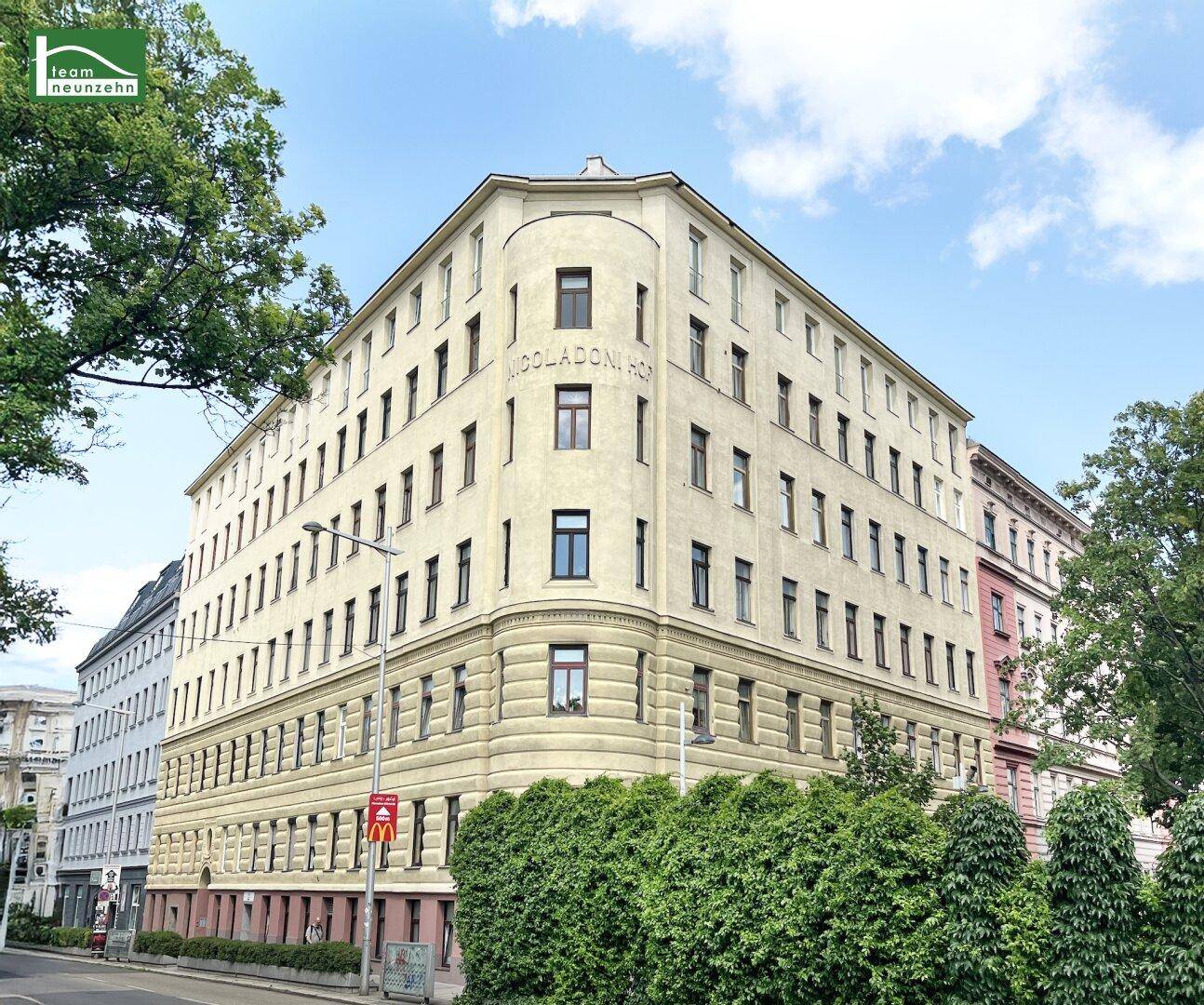 Elegantes Eckgebäude mit klassischer Architektur, vielen Fenstern und grünen Hecken an einem sonnigen Tag.