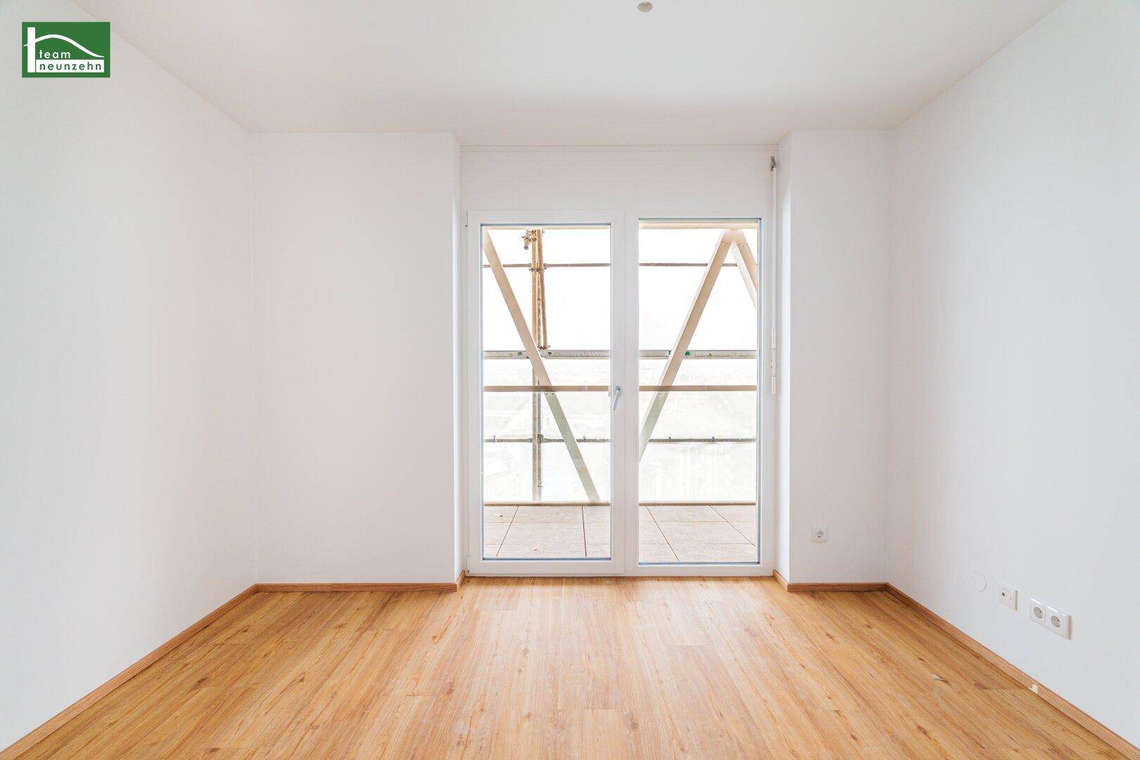 Heller Raum mit Laminatboden und großer Fenstertür, die viel Tageslicht hereinlässt.