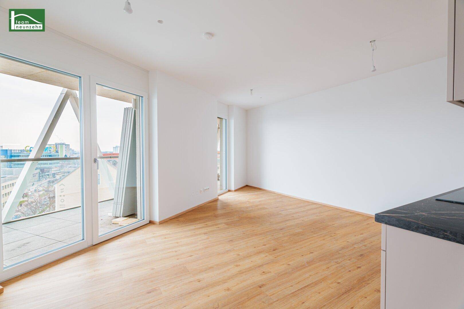 Heller, leerer Wohnbereich mit großen Fenstern und Zugang zu einem Balkon, ausgestattet mit Holzboden.