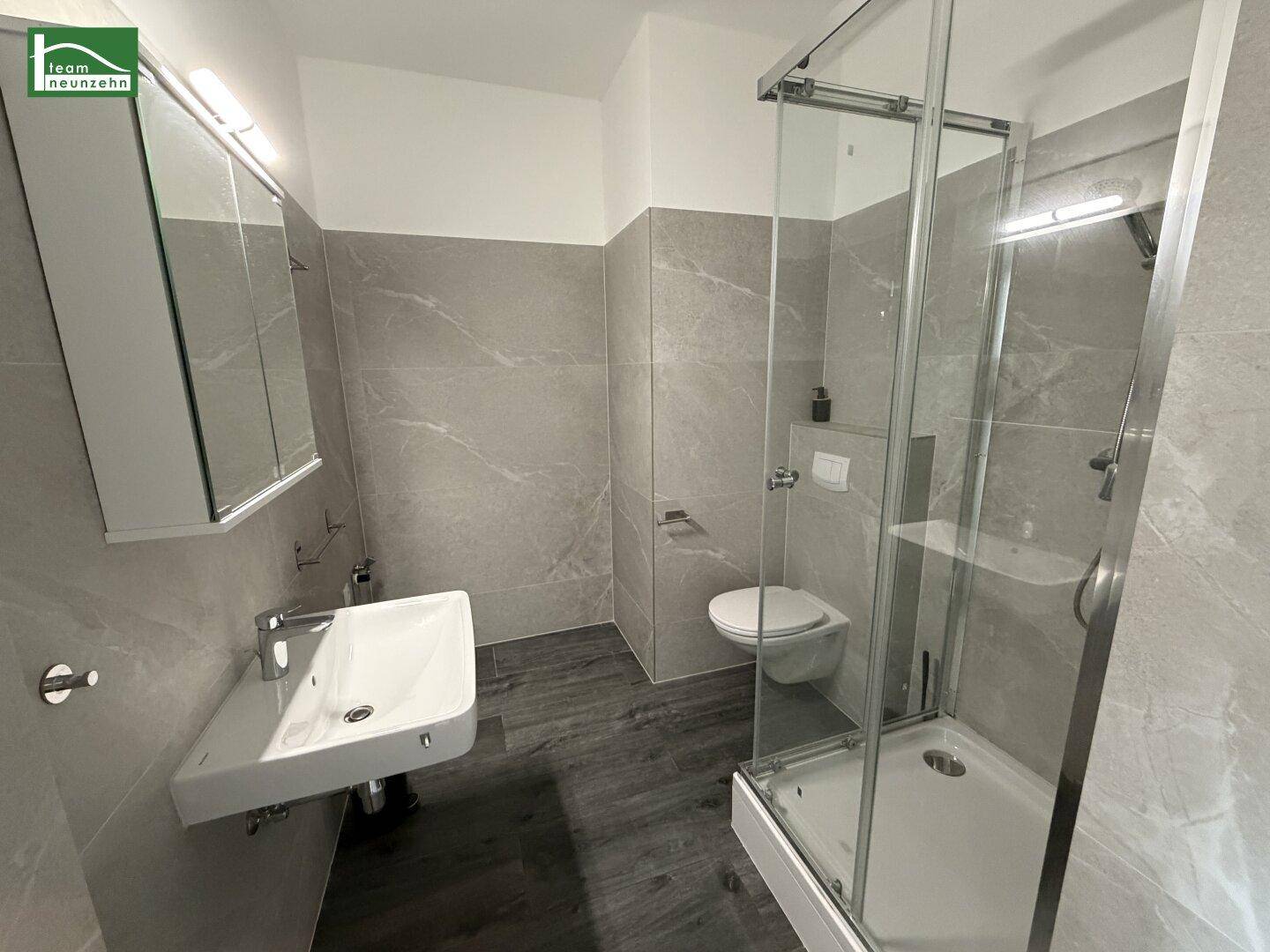 Modernes Badezimmer mit grauen Fliesen, Duschkabine, Waschbecken und wandhängendem WC.