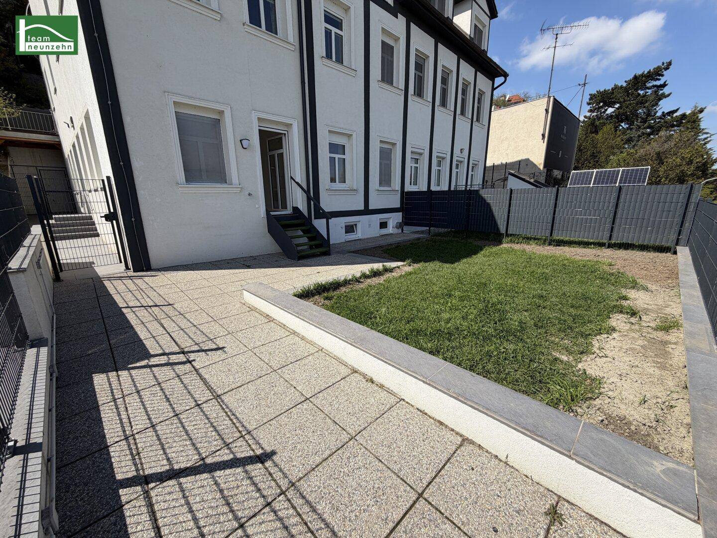 Außenbereich des Gebäudes mit gepflasterter Terrasse und Rasenfläche, umgeben von einem Zaun.