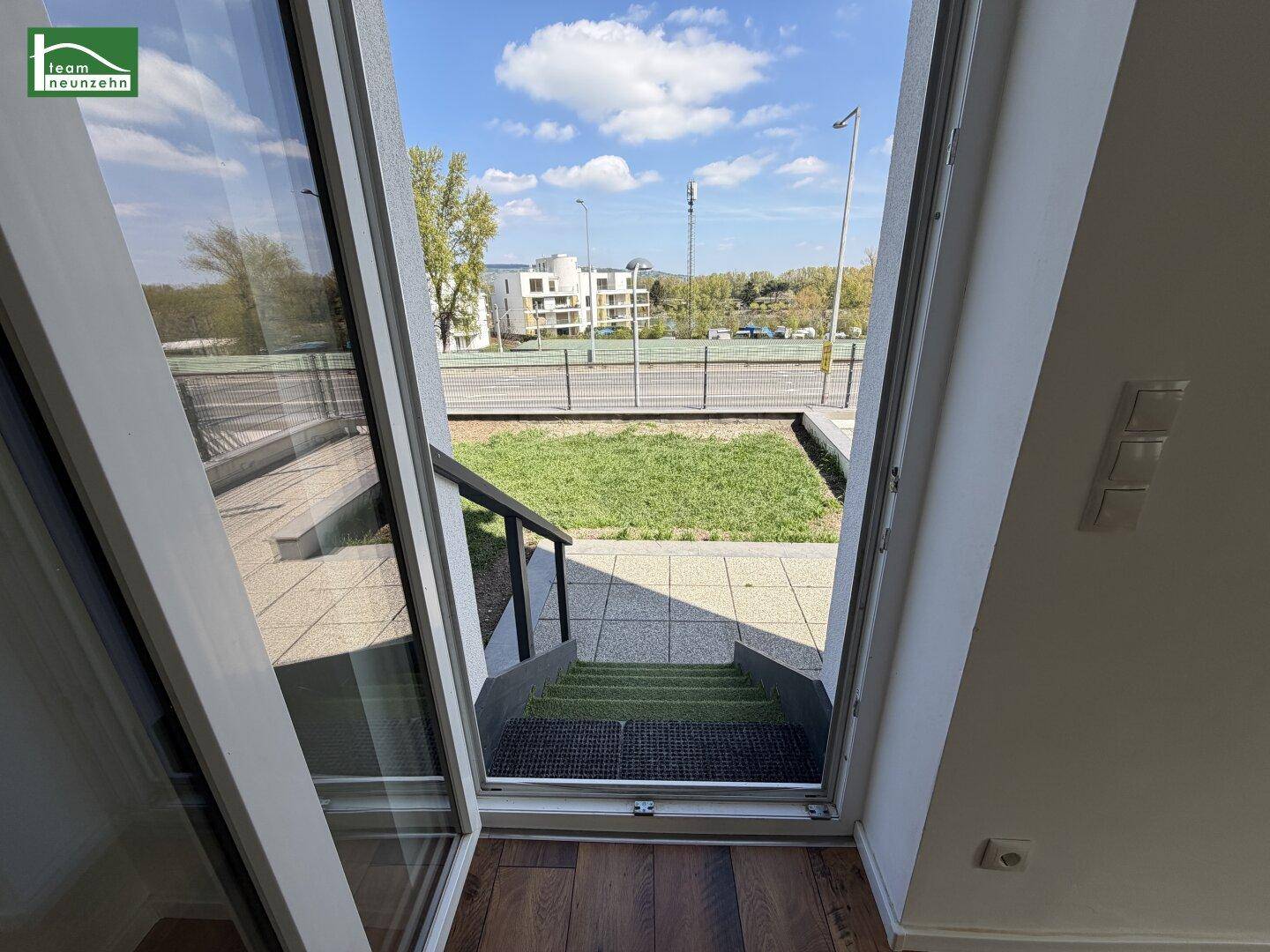 Blick aus dem Innenraum auf die Außenterrasse mit Grünfläche und umliegender Bebauung.