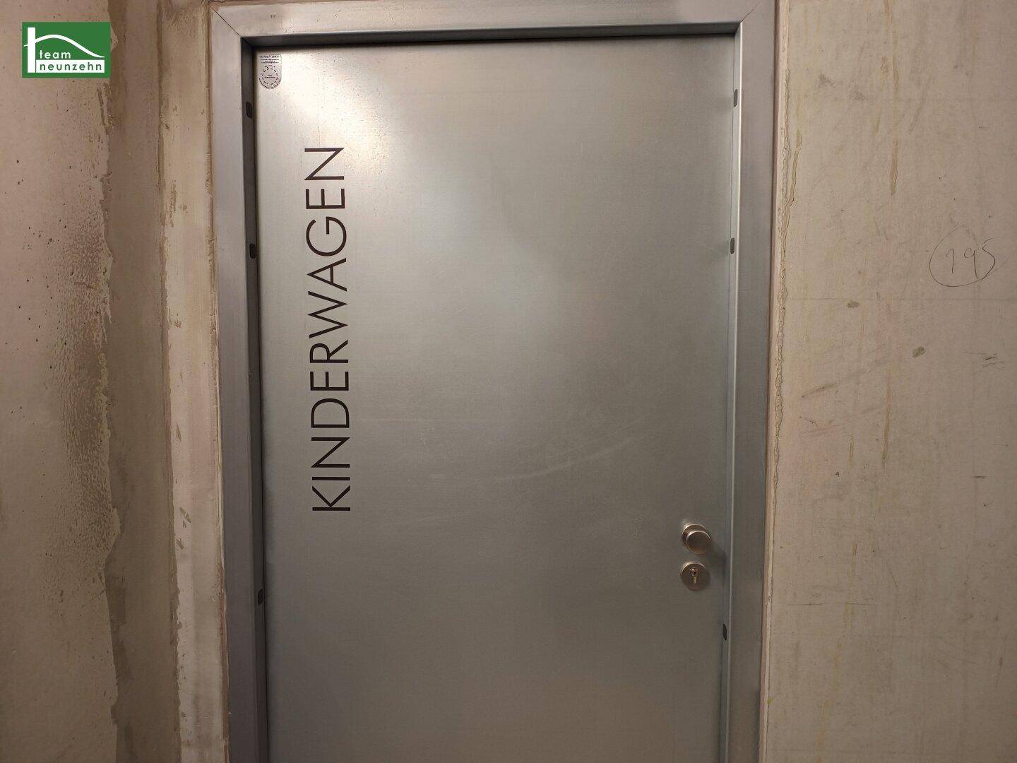 Eine Metalltür mit der Aufschrift "KINDERWAGEN" in einer Betonwand, für Kinderwagenabstellraum.