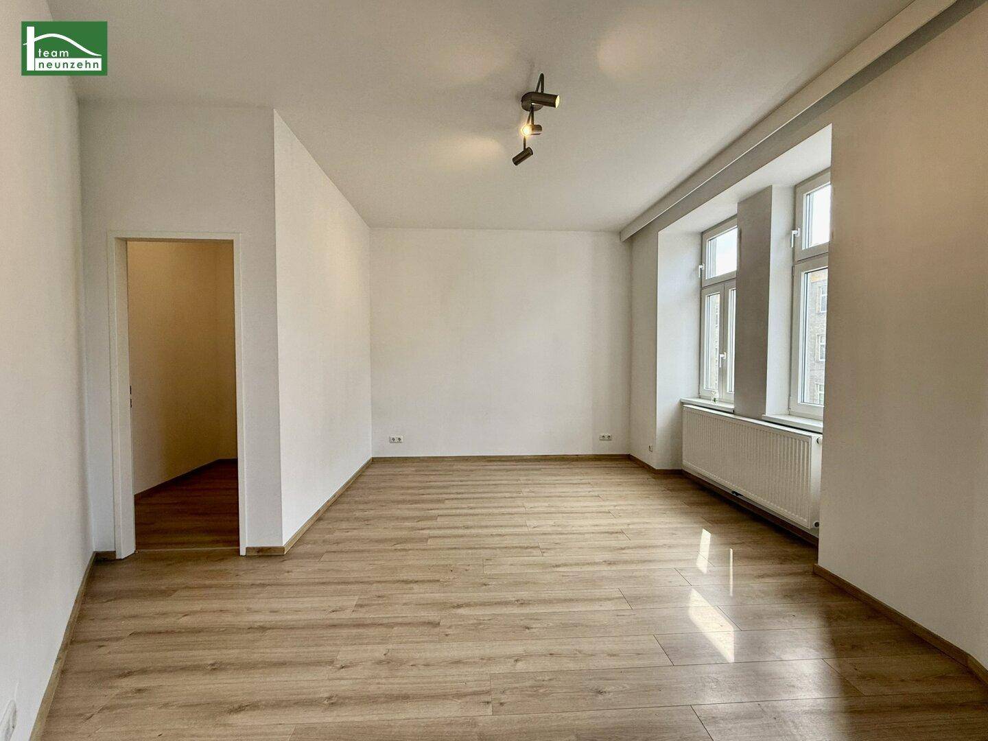 Geräumiges, unmöbliertes Wohnzimmer mit Laminatboden und zwei Fenstern für gute Belichtung.