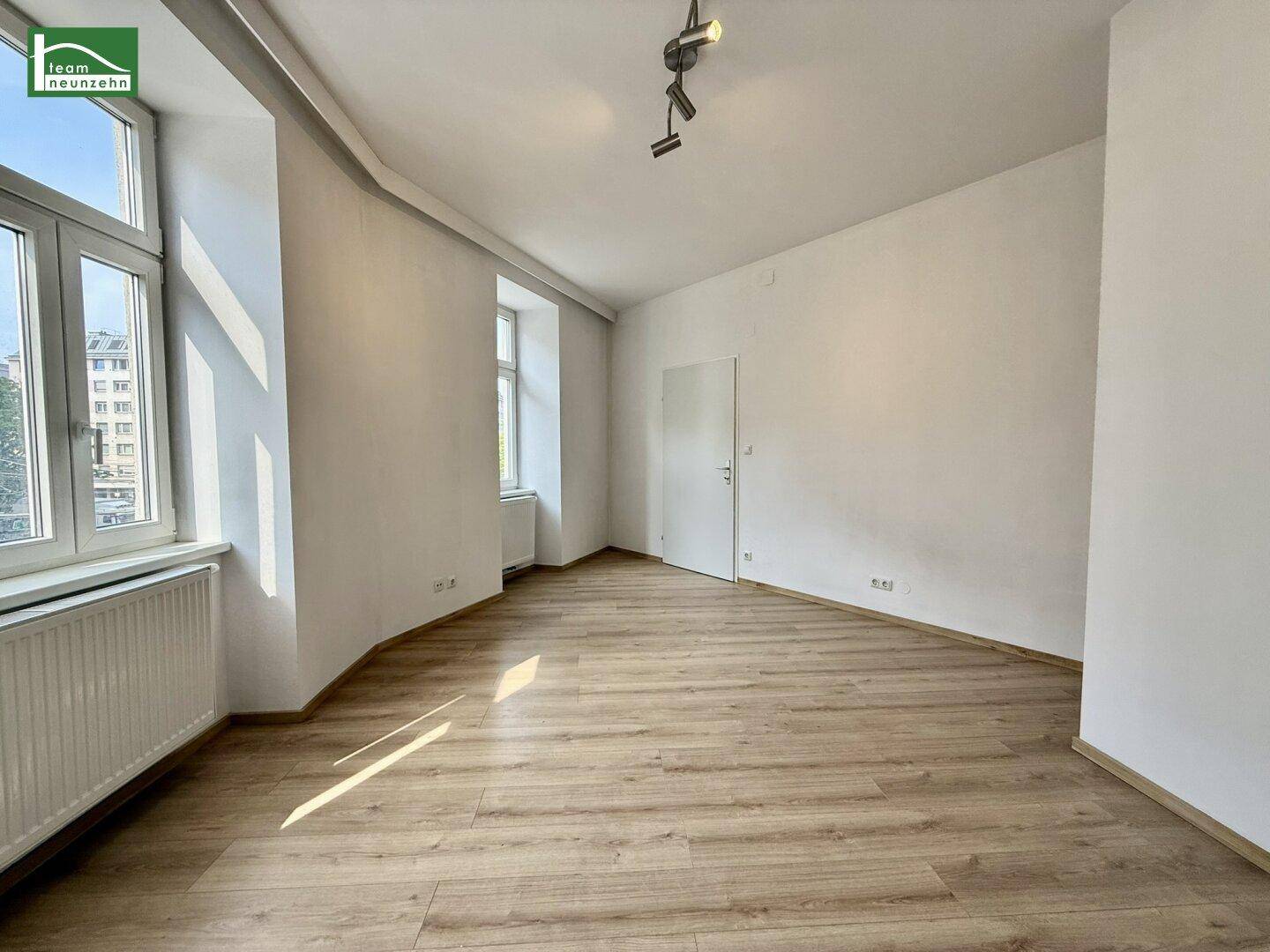 Leeres Wohnzimmer mit hellem Laminatboden und viel Tageslicht durch die großen Fenster.
