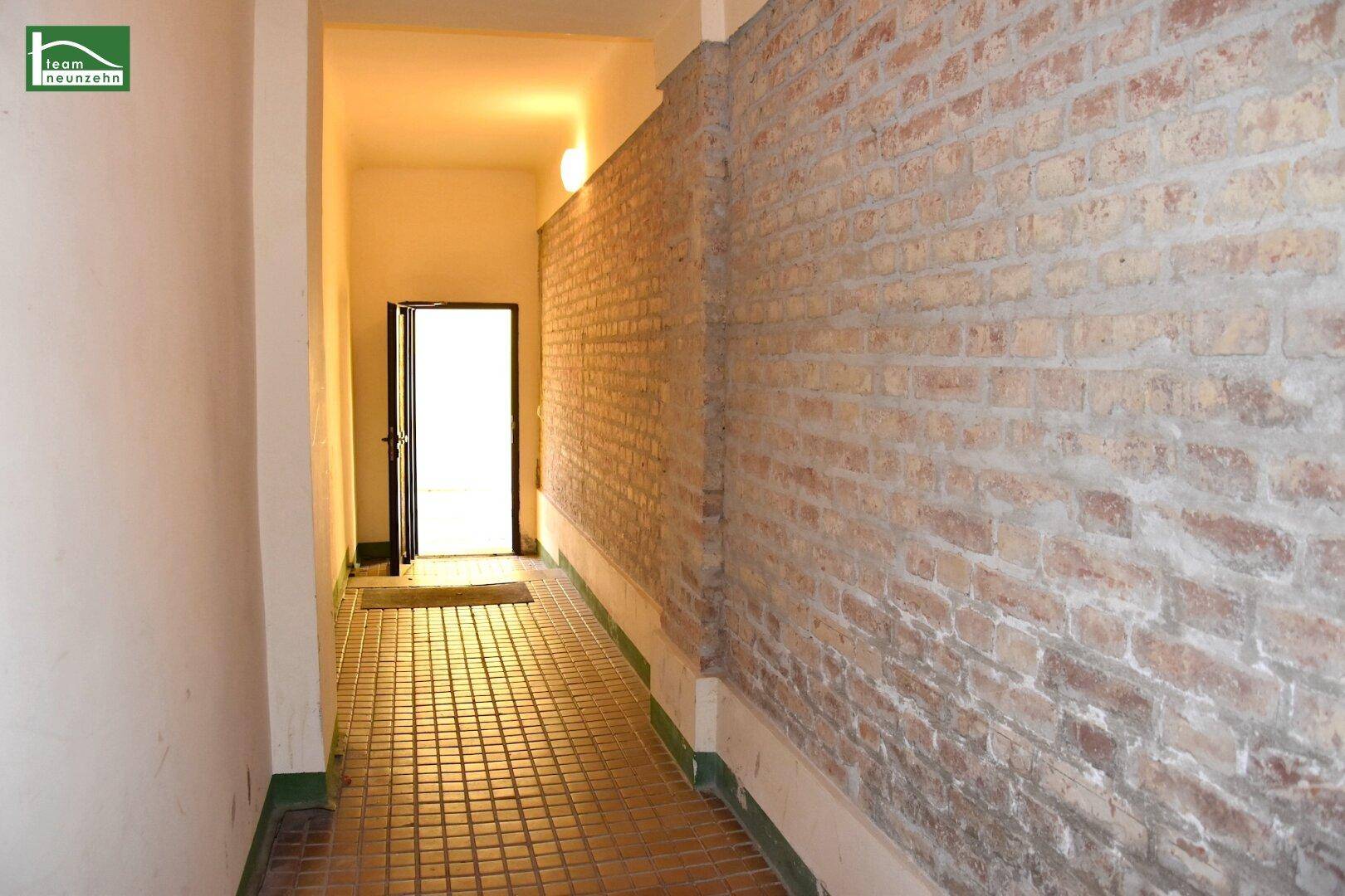 Langer Flur mit unverputzter Ziegelwand und gefliestem Boden, der zu einem hellen Ausgang führt.