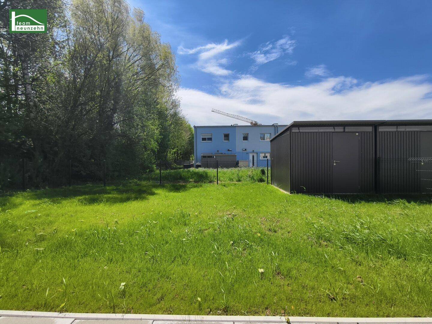 Grüne Rasenfläche mit dunklen Gartenhäusern und einem blauen Gebäude im Hintergrund unter klarem Himmel.