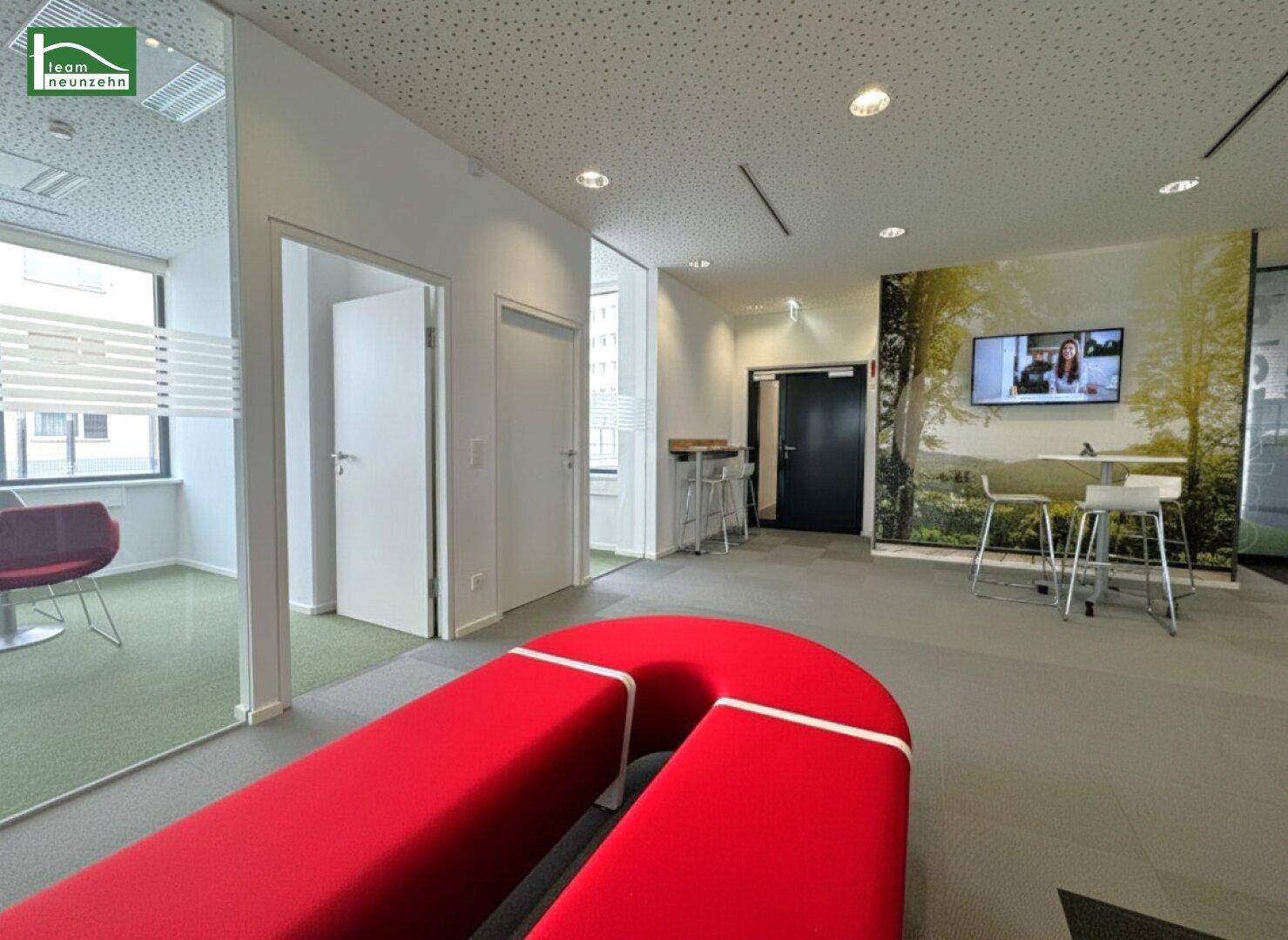 Moderner Loungebereich mit roten Sitzgelegenheiten, Glaswänden und einem Bildschirm für Präsentationen.